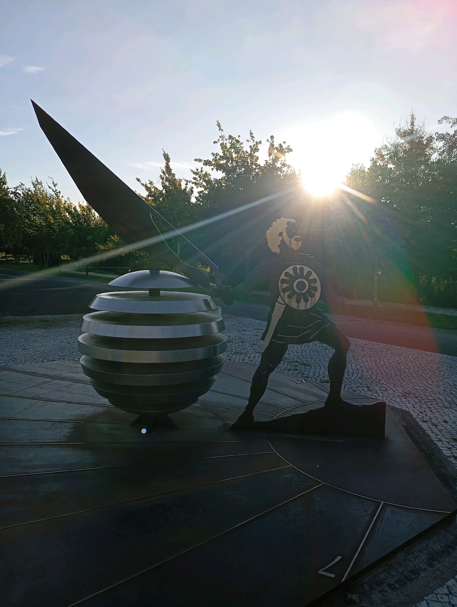 The image shows a sculpture outdoors, likely in a park or public space in Viseu, Portugal, given the provided context. The sculpture features a human-like figure with a shield, standing next to a large, layered spherical object with a pointed element extending upwards. The sun is setting or rising, casting a bright glare and lens flare across the scene, partially obscuring the upper portion of the sculpture and the sky. Trees and foliage are visible in the background, and the ground is paved with cobblestones and a dark, flat surface with geometric divisions. The overall atmosphere is serene, with natural light dominating the composition.