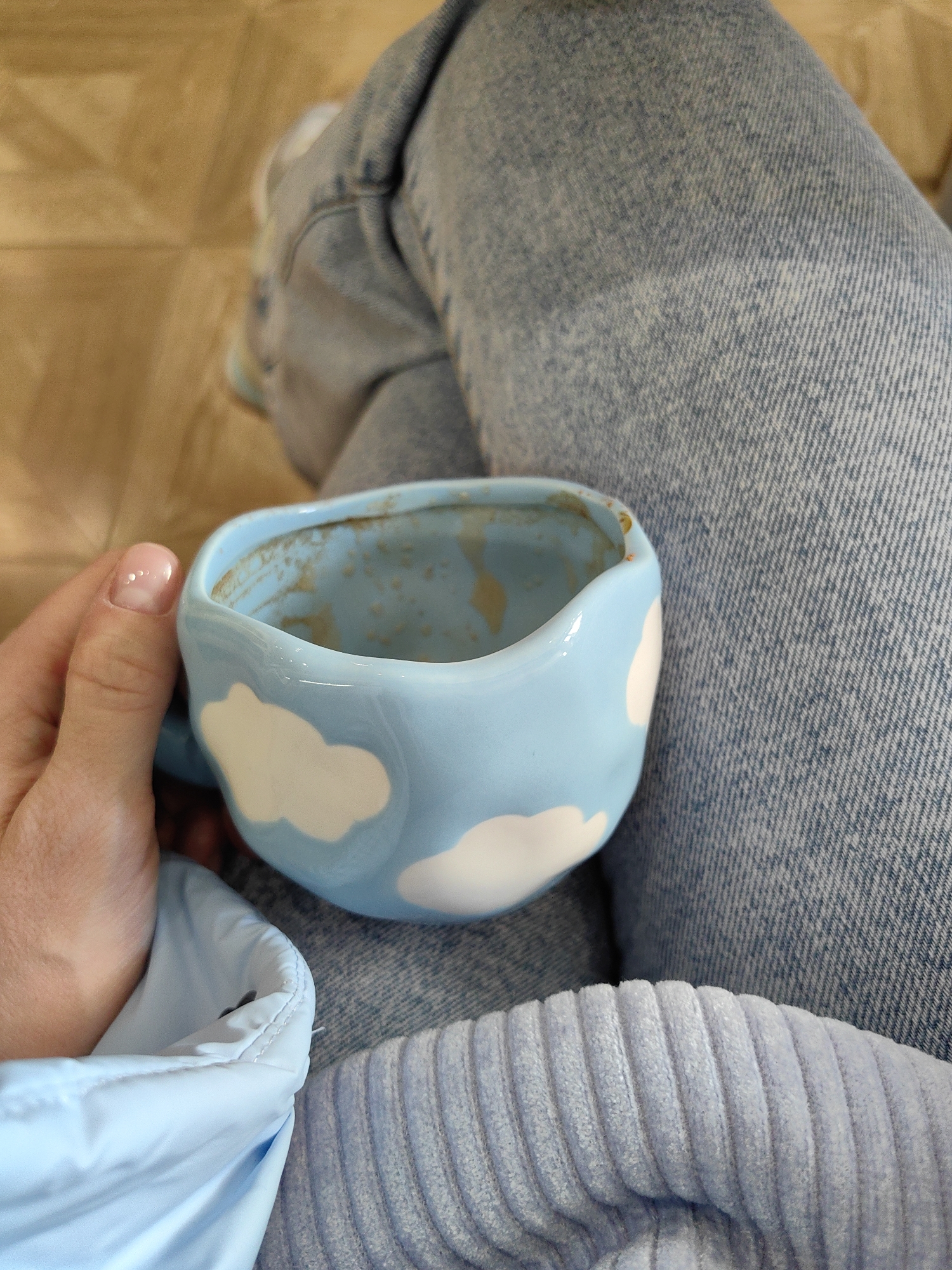 A person is holding a light blue, irregularly-shaped ceramic mug with white cloud designs in their left hand. The mug appears to be partially empty with some residue inside. The person is wearing blue jeans and a light blue corduroy shirt. The person's legs are crossed. The background is a light brown tiled floor.
There is no information in the image that identifies the location as Domodedovo, Russia.