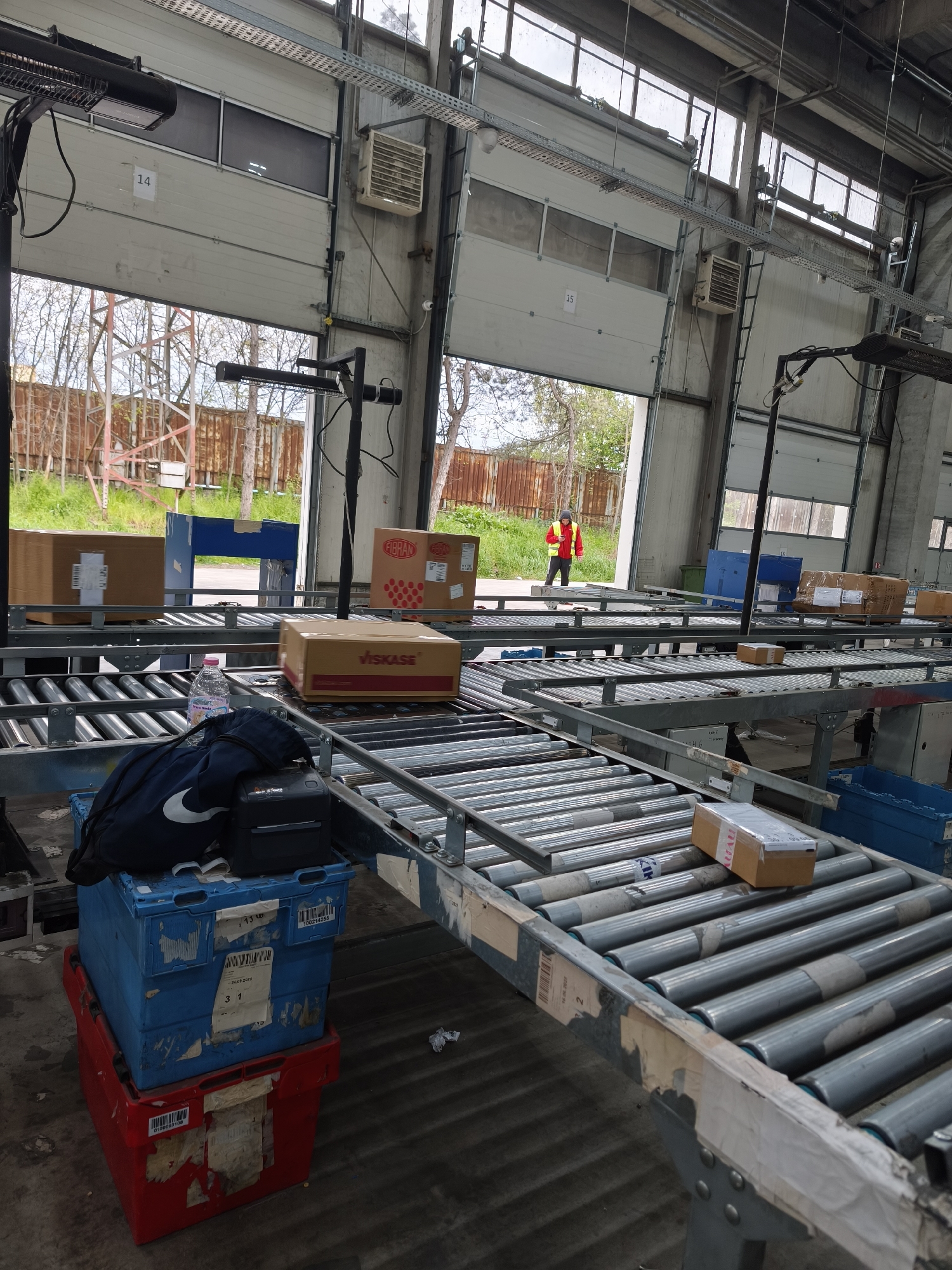 The image captures an interior view of an industrial loading dock or warehouse in Burgas, Bulgaria, bathed in natural daylight from multiple open bays and upper windows. The scene is dominated by an extensive system of metal roller conveyors, actively used for handling packages.
Several cardboard boxes are visible on and around the conveyors. Notable labels include "FIBRAN" on a larger box and "VISKASE" on another, while a smaller box partially shows "IMAU." Various shipping labels and barcodes are present on the packages. In the foreground, personal items rest on a blue plastic crate, which is stacked on a red crate. These items include a black backpack featuring a white Nike logo and a small black printer or device. A plastic bottle of "Gorna Banya" water is also on the conveyor. Visible text on the blue crate includes a date "24.08.2023" and numbers "3 1," while the red crate has a barcode starting with "0100504108."
Beyond the open loading bays, an outdoor environment with trees, green foliage, and a rusty metal fence is visible. A person in a red jacket and a high-visibility yellow vest is seen walking outdoors, away from one of the bays. The large white garage doors are numbered "14" and "15." Above the bays, industrial heaters or air handlers are mounted, and metal cable trays run across the ceiling. The setting conveys a functional and busy logistics operation, likely involved in package sorting or distribution.