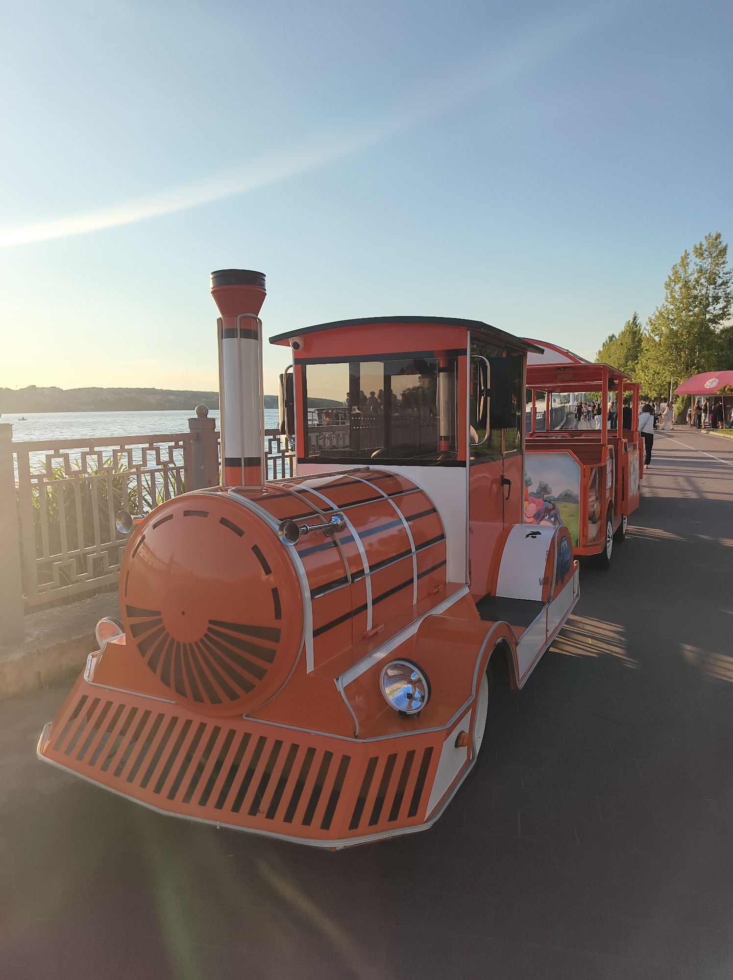 An orange and white toy train is parked on a paved pathway next to a body of water. The train has a red roof and white windows. Several people are visible walking in the background along the pathway, and a red tent is set up on the right side of the image. The sky is clear and blue with a few wispy clouds. The train appears to be a tourist attraction. The location is the Ternopil Lake promenade, Ternopil, Ukraine.
