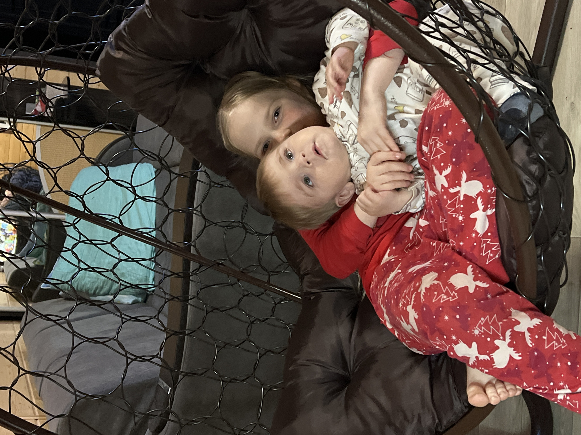 Two Caucasian children, one older girl and a younger toddler, are sitting in a brown, cushioned hanging chair within a room. The toddler is wearing red and white holiday-themed pajamas and is cradled in the girl's arms. The girl is wearing pajamas with a cream-colored base and a cartoon animal pattern. The chair has a dark metal frame and a wire-like backing. In the background, another chair is visible, along with what appears to be a light blue pillow. The exact location within the city of Bol'shakino, Russia, is indeterminable.