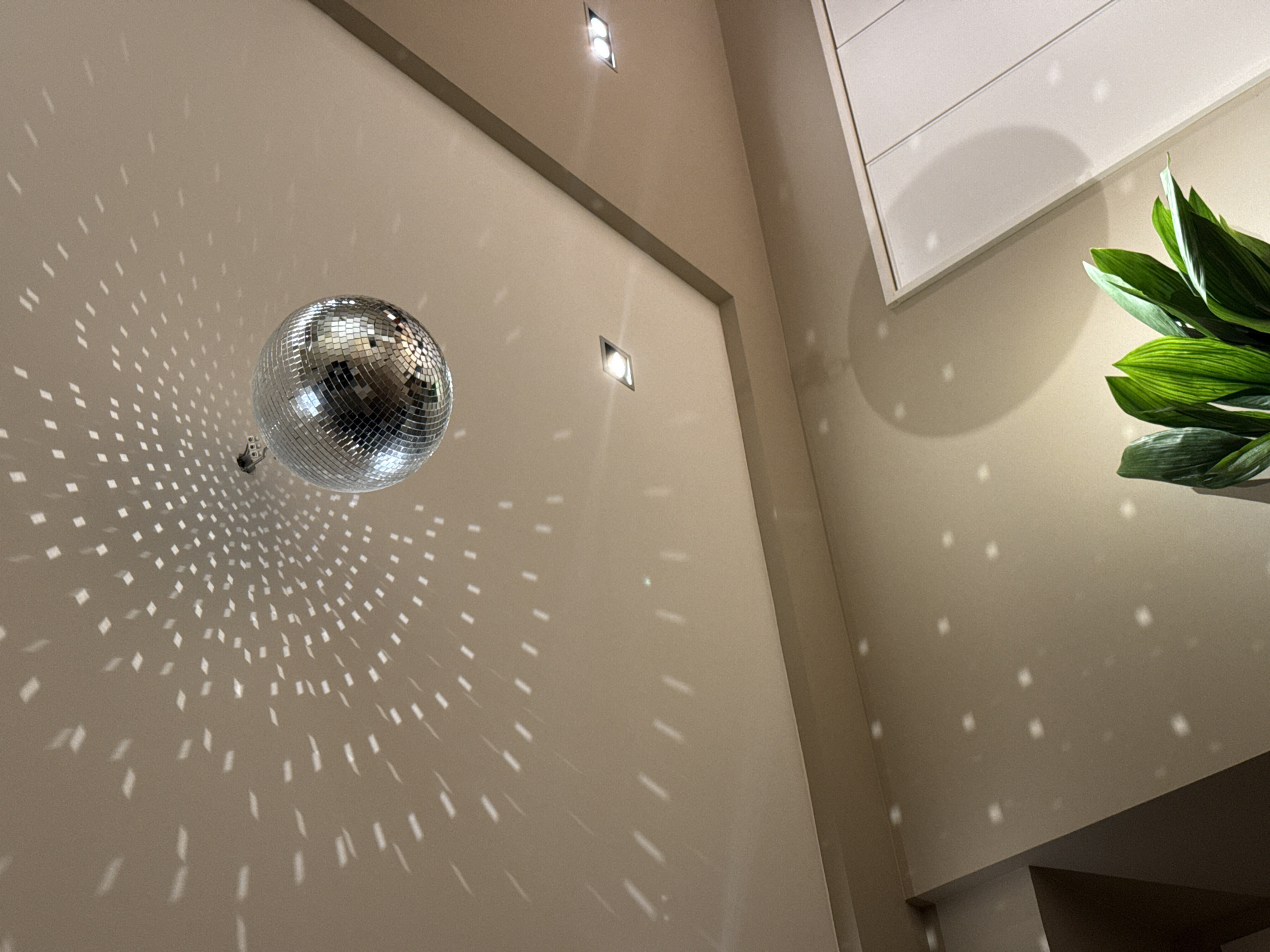 The image shows an interior scene featuring a disco ball suspended near the corner of a wall, reflecting light onto the surrounding surfaces in a pattern of small squares. The walls are light beige and feature recessed lighting. Part of a plant with large green leaves is visible in the upper right of the frame. The location cannot be determined from the image. There are no people visible.