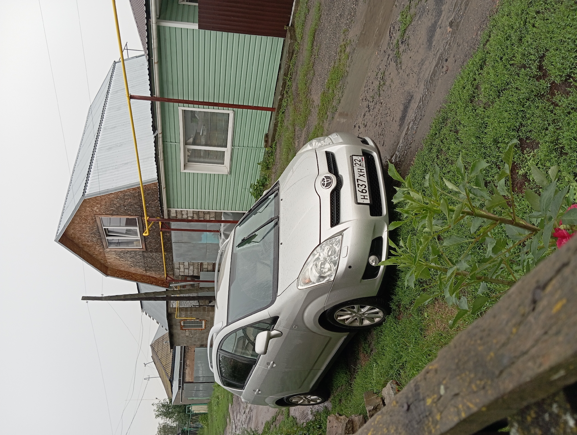 A silver Toyota Corolla Verso is parked on a muddy road next to a green vinyl-sided house. The house has a large wooden-gabled roof extension. A wooden fence is in the foreground. The license plate on the car reads "H 637 XH 22 RUS." The sky is overcast and the ground is wet, indicating recent rain. The scene appears to be in a rural or suburban area, consistent with the outskirts of Barnaul, Russia.