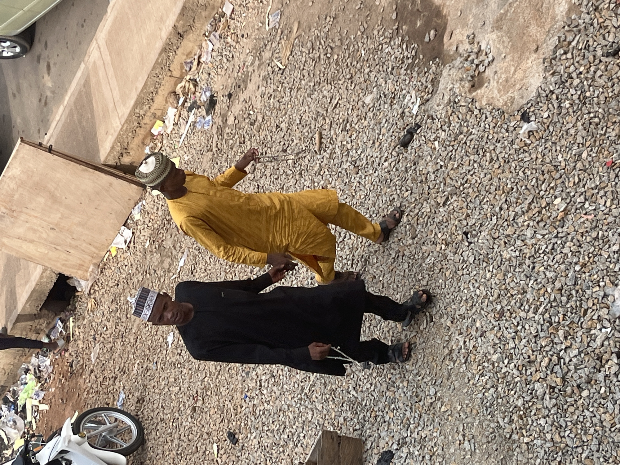 In the city of Jos, Nigeria, two young men are walking side-by-side on a gravel path, which appears to be part of a street. The man on the left is wearing a black traditional outfit and a patterned cap. He is holding a string of beads in his right hand and is looking towards the left. The man on the right is wearing a yellow traditional outfit and a cap. He is holding a string of beads in his right hand and is looking towards the right. Behind them, there is a pile of trash and debris, and a white motorcycle is parked on the left. The ground is covered in small grey and brown gravel.