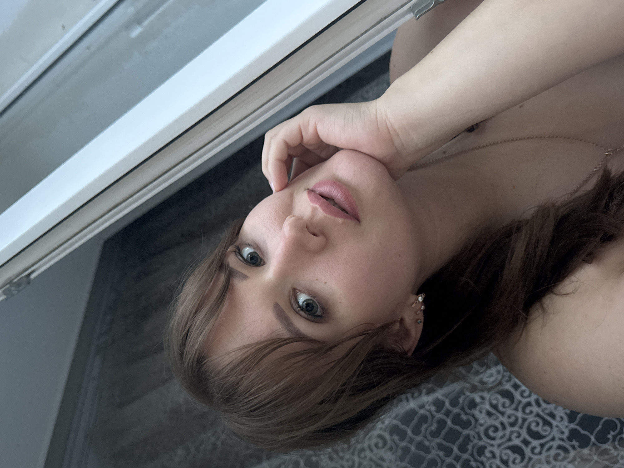 A young woman with light brown hair and bangs is posing for a selfie indoors, near a window. She is resting her chin on her hand and looking at the camera. She is wearing earrings and a necklace. The background consists of a curtain and a window frame. The image is cropped to focus on her face and upper body. The location is Chaikovsky, Russia.