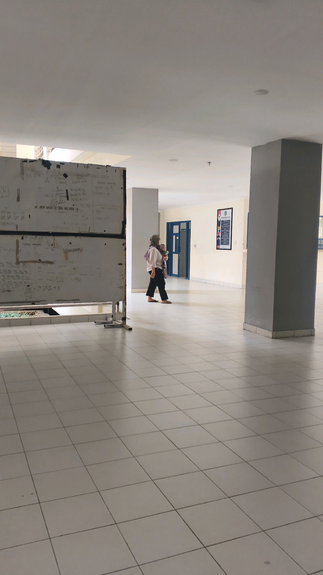 A full-shot vertical-oriented, eye-level image captures an indoor tiled hallway with a woman carrying a baby. The hallway is well-lit with fluorescent ceiling lights and natural light from openings above. To the left, a large white board covered in handwritten notes and equations stands on a metal frame. The woman, wearing a light-colored shirt, black pants, and a hijab, walks to the right, carrying a baby strapped to her chest. She appears to be mid-stride. In the background, a blue double door is visible, as well as a framed poster on a cream-colored wall. A large grey rectangular column is on the right side of the frame. The floor is made of light-colored square tiles laid in a grid pattern. The scene is devoid of outdoor elements, suggesting an indoor public space like a school or office building. There are no discernible landmarks or street signs that can definitively place the image in Lohbener, Indonesia, but the setting is consistent with architectural styles found in the region.