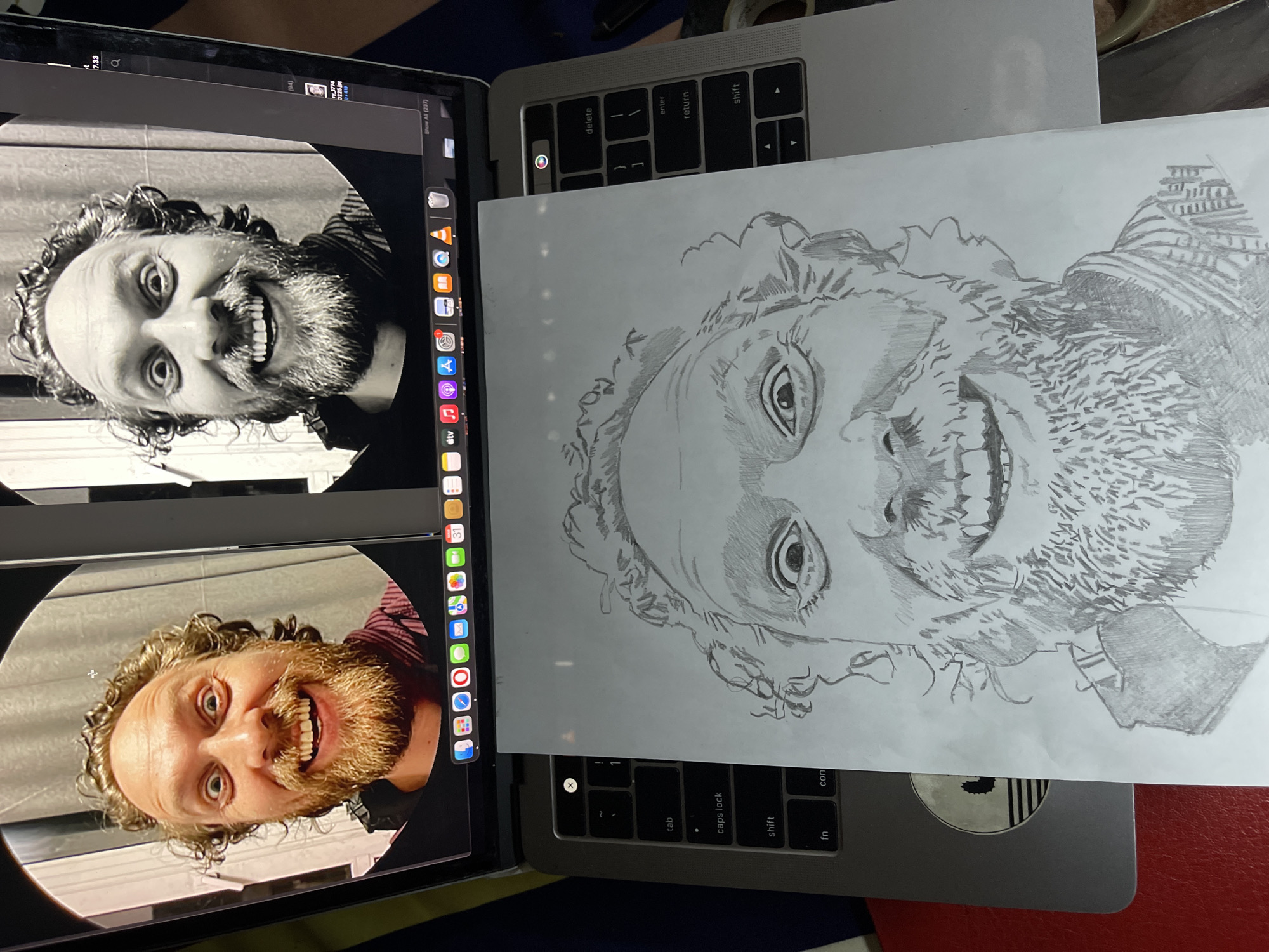 A close-up, top-down shot displays two laptops and a drawing. The laptop on the left, an iPad, has two images of a bearded man with curly hair, grinning widely. The upper image is black and white, while the lower one is in color. The iPad's dock with various application icons is visible on the right edge of the screen.
The laptop on the right is a MacBook, which is partially covered by a white sheet of paper with a pencil sketch of the same man. The sketch is incomplete but includes the man's face, hair, and beard. Parts of the MacBook keyboard are visible around the paper.
The overall composition presents a comparison between the original images on the iPad and the drawn representation on paper. The scene gives no indication of Jos, Nigeria.