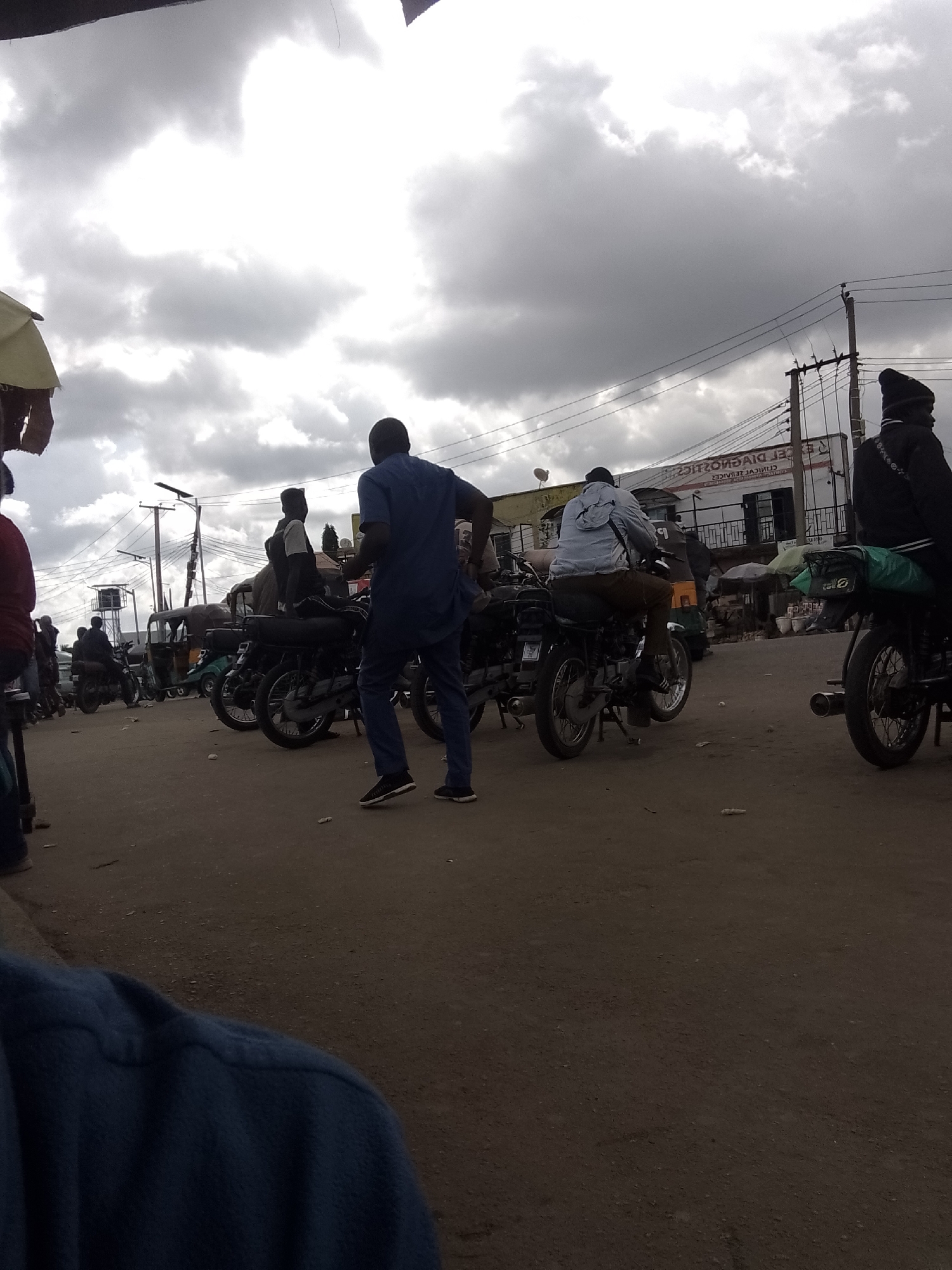 In Gwong, Nigeria, a street scene unfolds under a cloudy sky. Several individuals are present, some on motorcycles and others standing. One person, wearing a blue dashiki and pants, stands in the foreground, facing away from the camera. Another person in a light-colored jacket and brown pants is seated on a motorcycle, moving away from the viewer. A third person in a black jacket and beanie is also on a motorcycle, facing left. In the background, a building with a sign displaying "CLINICAL DIAGNOSTICS" is visible. The ground is dirt, and several motorcycles and a tricycle are parked or in motion. The overall atmosphere appears to be that of a busy marketplace or transport hub.