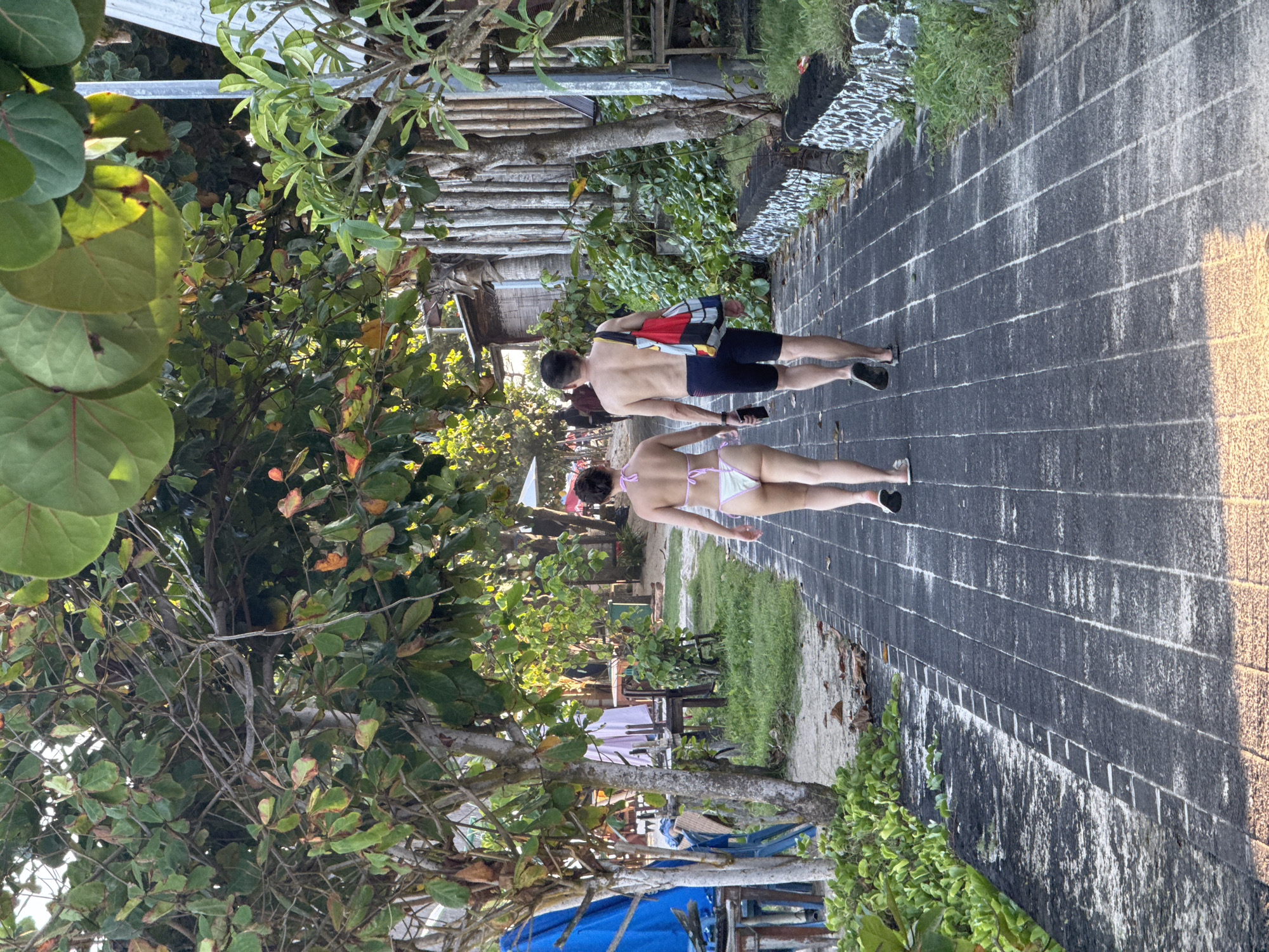 Two people, a man and a woman, are walking on a brick path. The man is shirtless and wearing dark-colored shorts, carrying a colorful bag over his shoulder. The woman is wearing a white bikini. The path is lined with greenery and trees. In the background, there are buildings, trees, and furniture. The location is Kuta Selatan, Indonesia.
