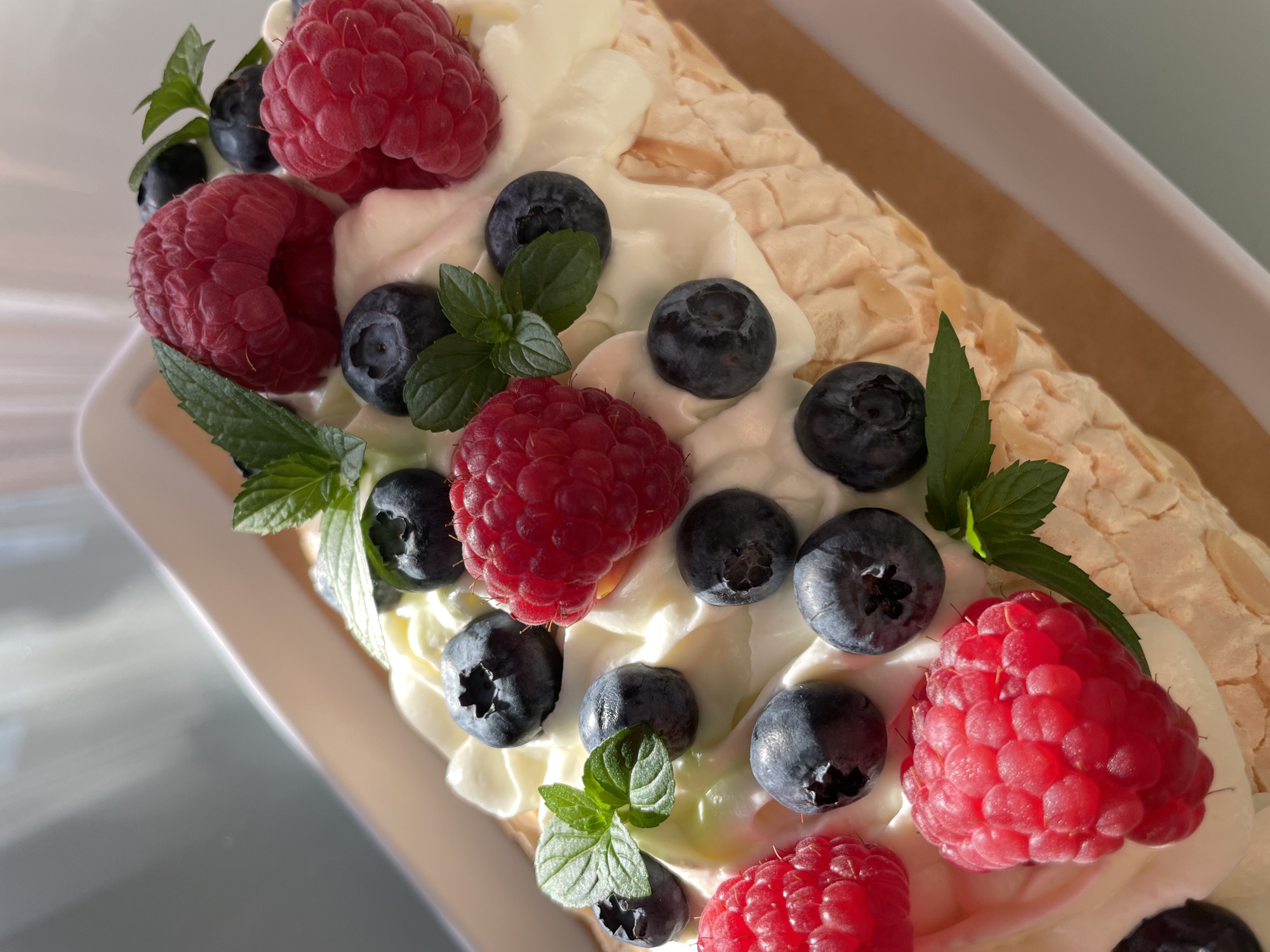 The image shows a close-up of a rectangular pavlova dessert topped with whipped cream, fresh raspberries, blueberries, and mint leaves. The dessert is served in a white rectangular dish, which is placed on a light brown surface. The lighting suggests it is daytime, possibly near a window, as evidenced by the bright light and shadows. The background is blurred and shows a light-colored, angled surface, likely a wall or table, with a hint of a grey-blue object. There are no people or animals visible in the image. The scene is entirely focused on the food item. There is no indication of this scene being located in Saint Petersburg, Russia.
