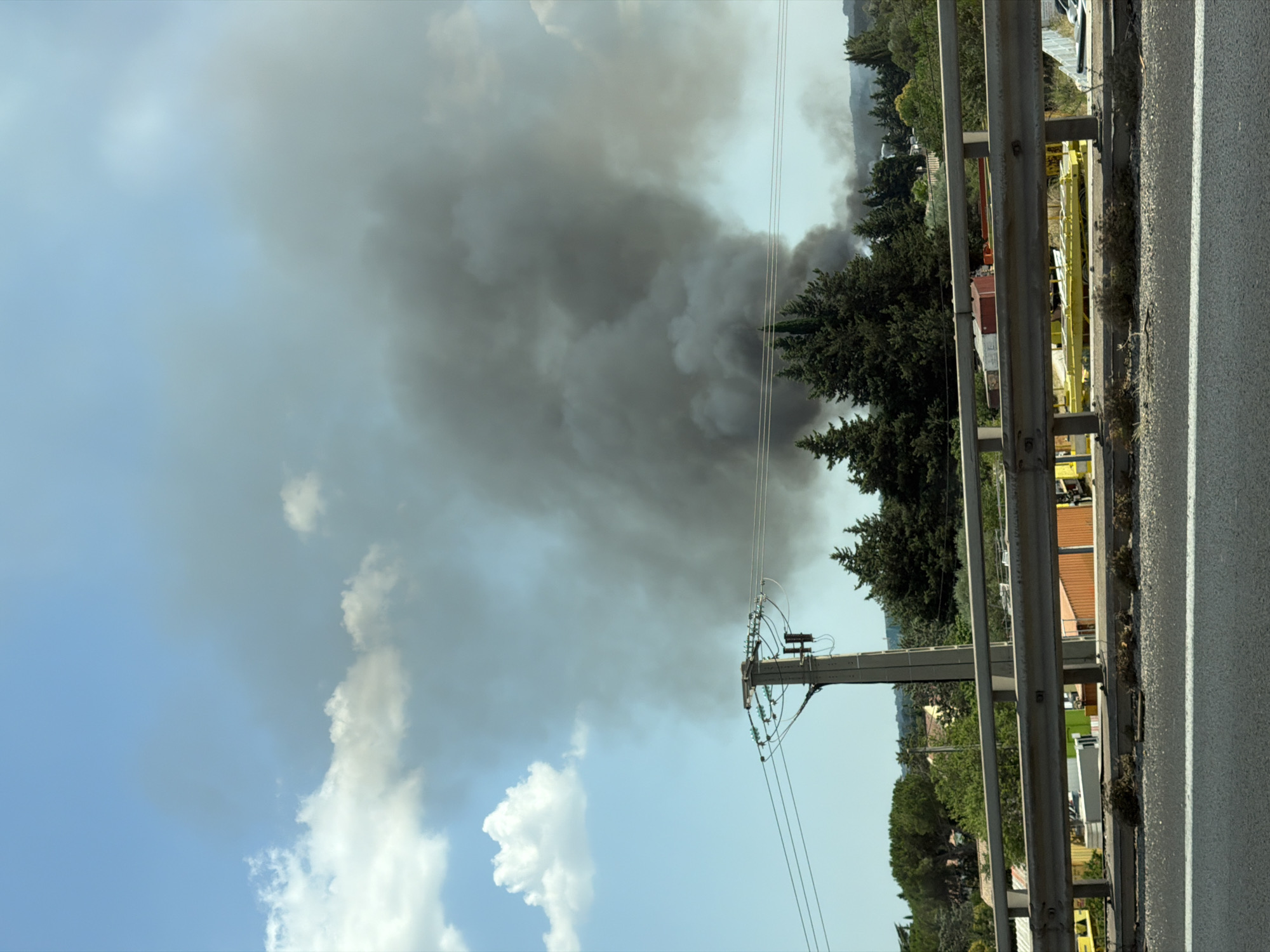 This is a vertically oriented, eye-level, wide-angle photograph taken outdoors on a clear, sunny day. The scene depicts a large plume of dark smoke rising into the sky, indicating a fire. The smoke is a dominant element, obscuring the sky and a portion of the distant landscape. In the foreground, there is a metal guardrail along the side of a road, with a paved road to the right. To the left of the guardrail, a utility pole with wires is visible, along with lush green trees and some buildings in the distance, suggesting a semi-rural or suburban setting. The overall impression is one of an unfolding emergency.