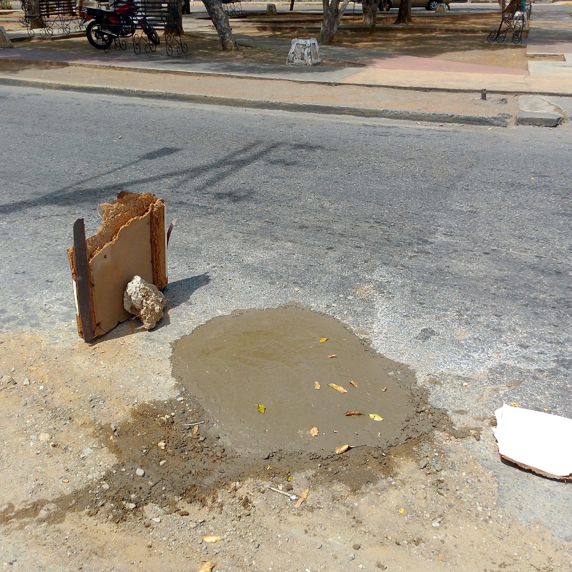 The image depicts a street scene in Altagracia, Venezuela. In the foreground, there's a puddle of muddy water on the asphalt, with scattered leaves. To the left of the puddle, there's a piece of damaged furniture or building material, consisting of a wooden frame with exposed interior padding and a rock propped against it. A piece of white material lies to the right of the puddle. The background shows a paved road leading to a sidewalk area with benches, trees, and a parked motorcycle.