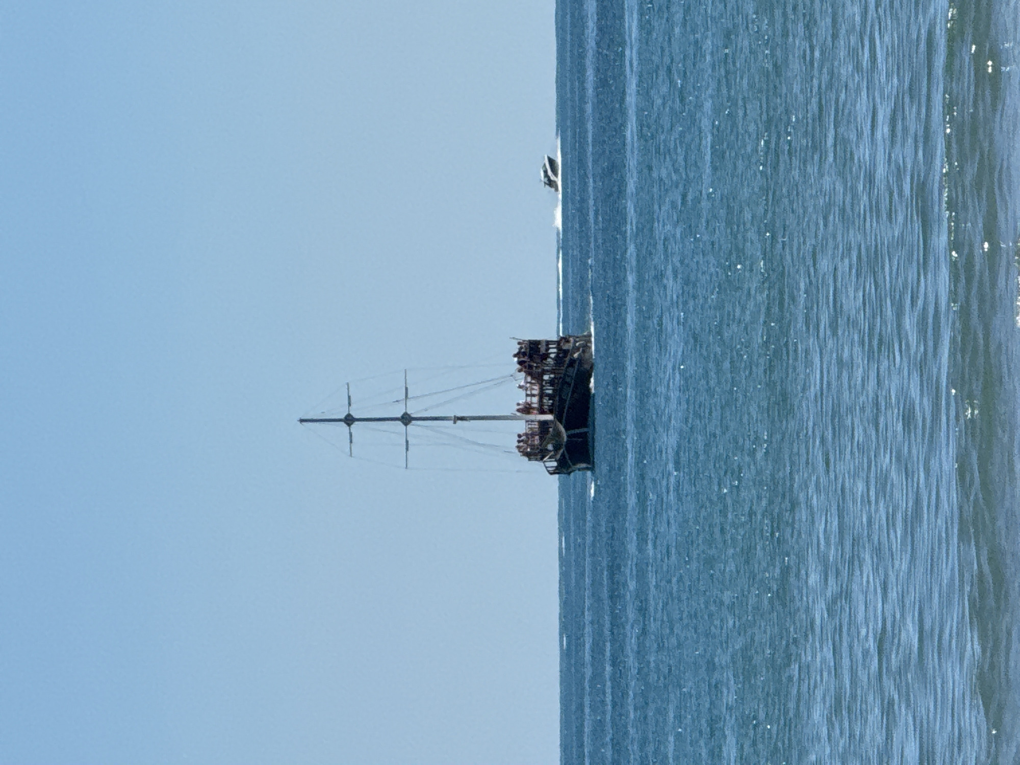 A pirate-style tourist boat with people on deck sails on the blue sea under a clear sky, with a smaller white motorboat in the distance. The photo was taken from the shore.