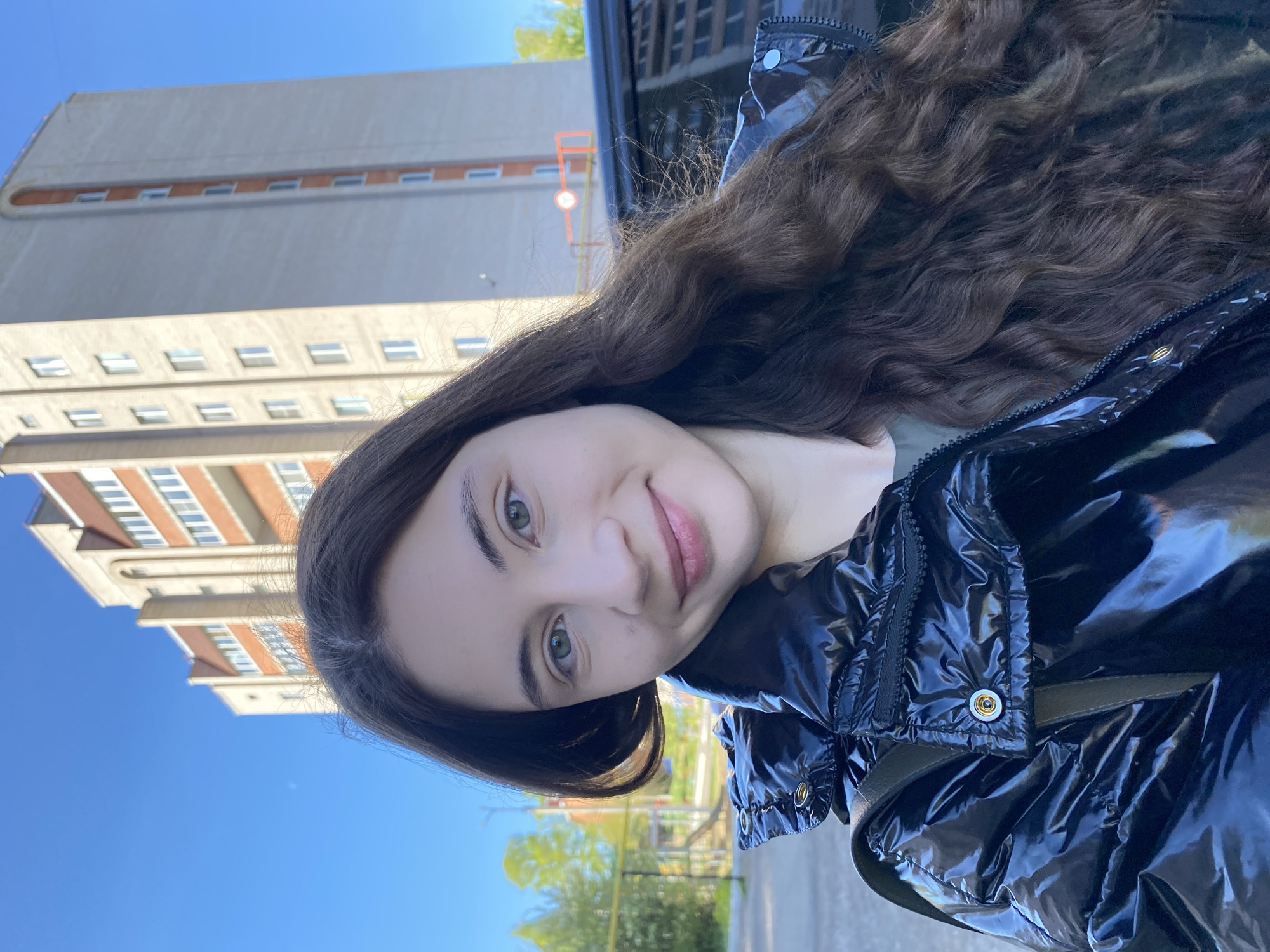 A young woman with long, wavy brown hair, light skin, and green eyes poses for a selfie outdoors. She is wearing a shiny black puffer jacket. Behind her is a tall, light-colored building with a series of windows, characteristic of Soviet-era architecture. The blue sky is visible above. The scene is set in Votkinsk, Russia, identified by the distinct architectural style of the building, typical of housing structures from that period.