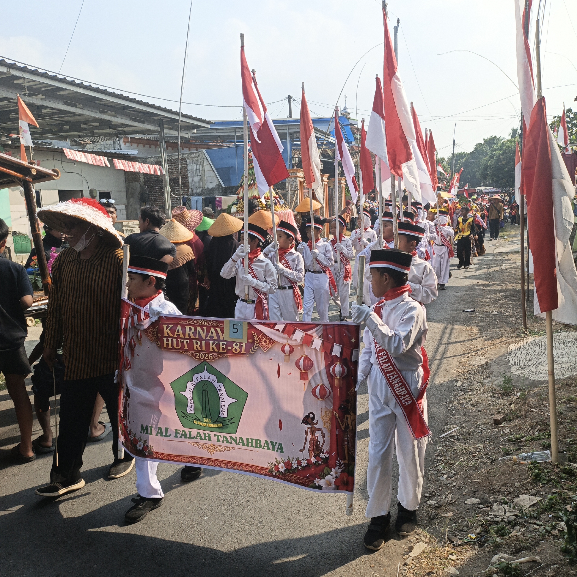 An outdoor daytime procession is occurring on a street in Tanahbaya, Indonesia. A group of children, dressed in white uniforms with red sashes and black and white hats, are marching in a line, carrying poles with red and white flags. They are holding a large banner that reads "KARNAVAL HUT RI KE-81 2026 MI AL FALAH TANAHBAYA" and features a green emblem with a candle and the text "YAYASAN AL FALAH TANAHBAYA". One child in the foreground is holding the banner.
To the left of the children, an adult man wearing a striped shirt and a large conical hat is also participating in the procession. Other adults and children are visible in the background, some wearing similar hats. The street appears to be paved with asphalt and has a dirt shoulder with some dry vegetation and debris. Buildings with corrugated metal roofs are visible along the side of the street. The sky is bright and clear, indicating sunny weather. The overall scene suggests a festive local event, likely a parade or carnival celebrating Indonesian Independence Day.