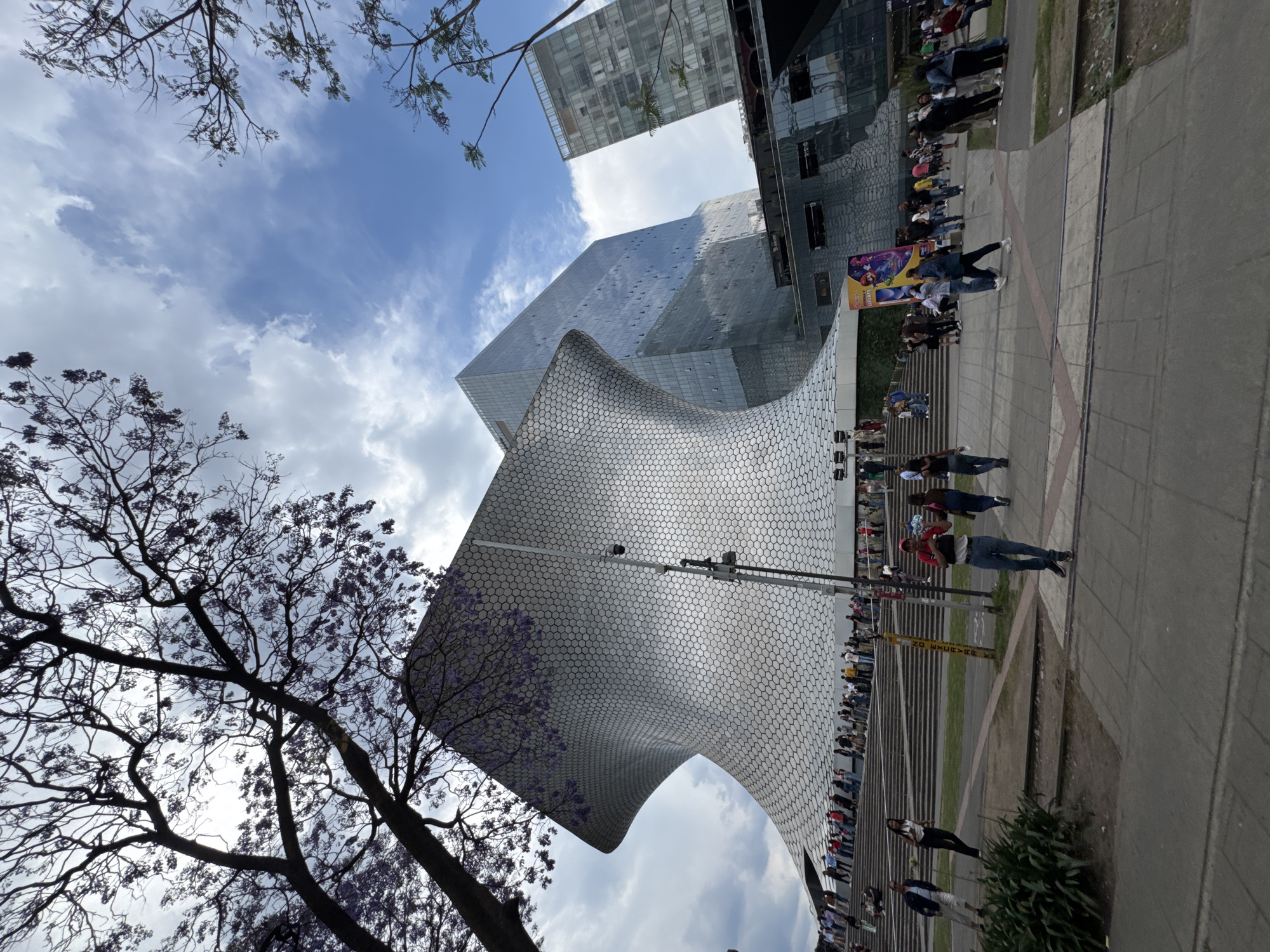 The image depicts the Soumaya Museum in Mexico City, Mexico, an iconic architectural structure characterized by its unique, curved, and reflective honeycomb-like exterior. A tree with purple flowers is in the foreground on the left. A line of people is seen in front of the museum. The sky is partly cloudy, and other tall buildings are visible in the background. The image appears to be taken from a low-angle perspective, emphasizing the museum's height and distinct design. People are walking around the area.
