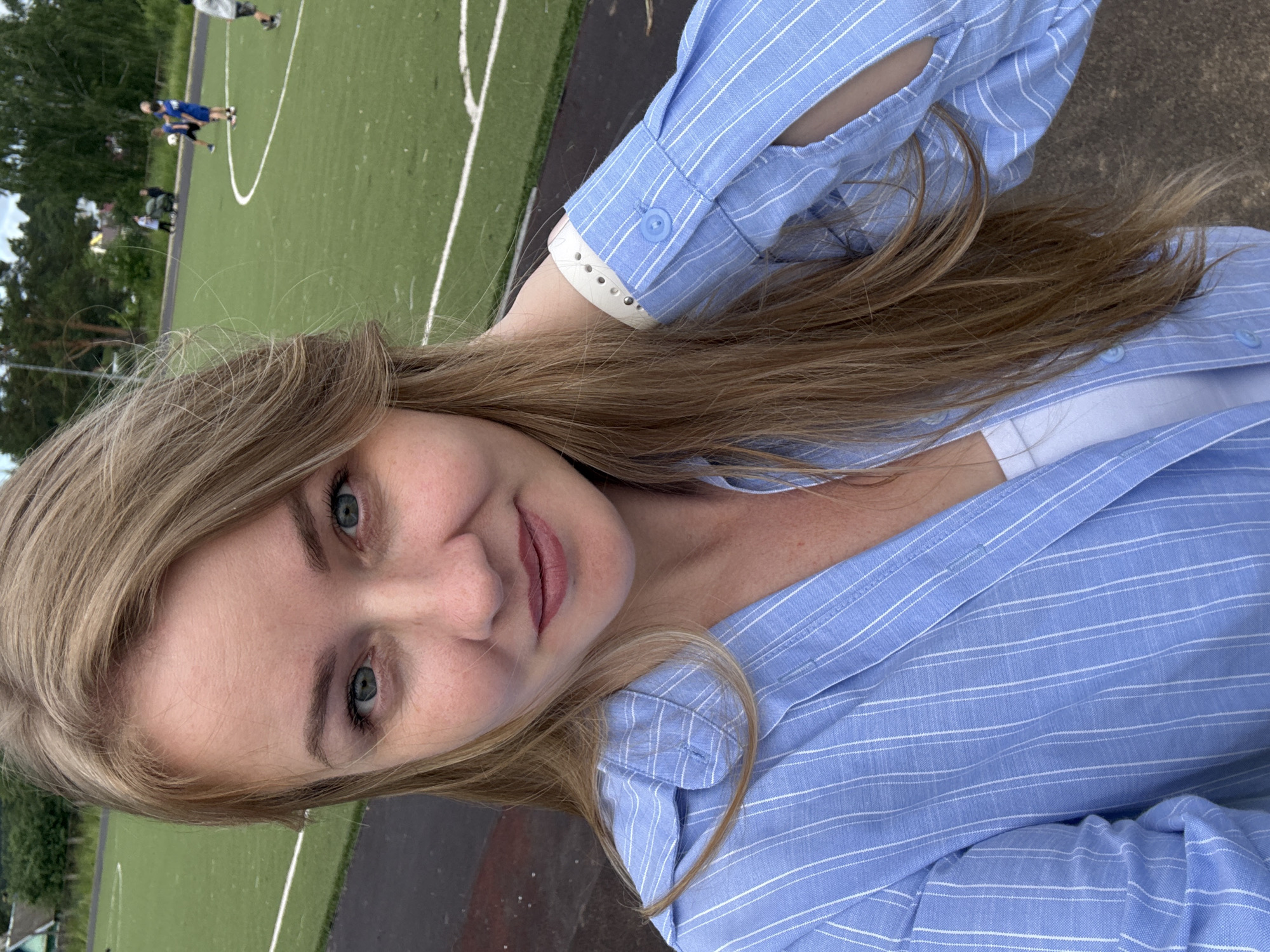 A blonde woman with blue eyes takes a selfie outdoors near a green sports field with white lines, possibly a soccer field. She is wearing a light blue and white striped button-down shirt over a white tank top and a light-colored bracelet on her wrist. In the background, several people, some appearing to be children, are on the field, with trees and buildings visible in the distance. The scene suggests a location within the city of Chaikovsky, Russia, potentially a local park or school sports complex.