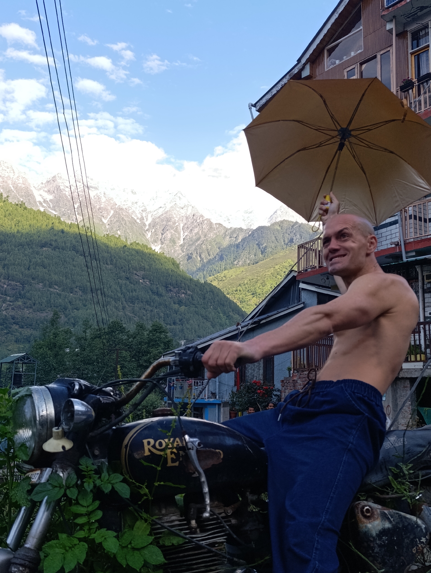 In Bahang, India, a shirtless, bald man with a tan is seated on a black Royal Enfield motorcycle that is partially overgrown with green foliage. He is holding a tan umbrella open above his head with his left hand. The man is wearing dark blue pants. He is looking to his right with a slight smile. The motorcycle is parked in front of a multi-story building with wooden siding and balconies. In the background, a lush green mountainside is visible, topped by snow-capped peaks under a partly cloudy blue sky. Power lines run diagonally across the upper left portion of the image.