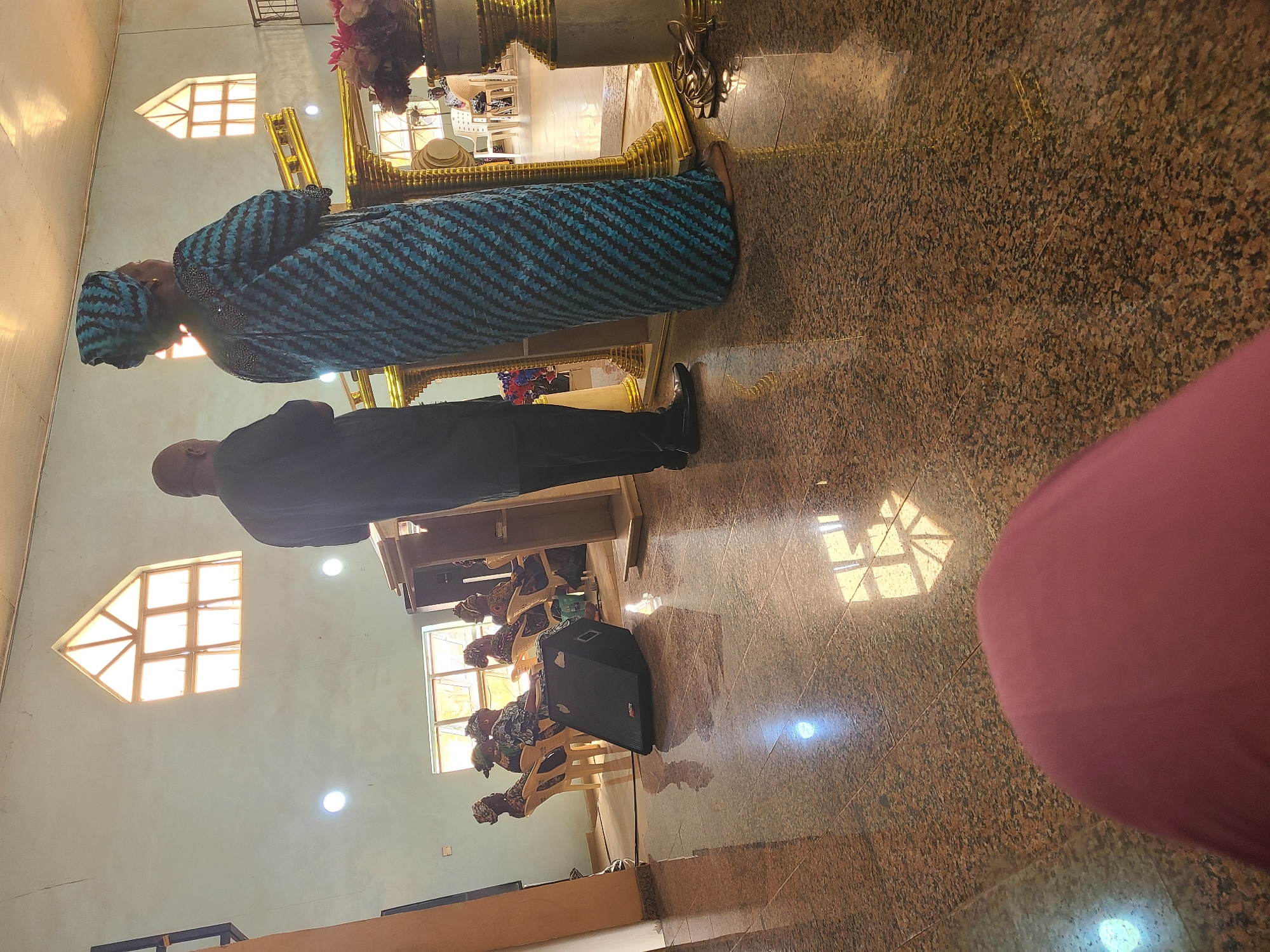The image depicts an indoor scene, likely a place of worship or a community hall, with polished granite floors reflecting light from windows and overhead fixtures. In the foreground, a person wearing a magenta garment is partially visible. Two individuals stand near a golden altar-like structure. The person on the left is a man in a dark suit and black shoes. The person on the right is a woman in a blue and black patterned dress and headscarf, facing away from the camera. In the background, several women are seated on light-colored chairs, also wearing headscarves and patterned clothing. A black speaker is positioned on the floor to the left of the seated individuals. Above, a triangular window with multiple panes is visible on a light blue wall, alongside circular lights. The overall ambiance suggests a gathering in Karu, Nigeria.