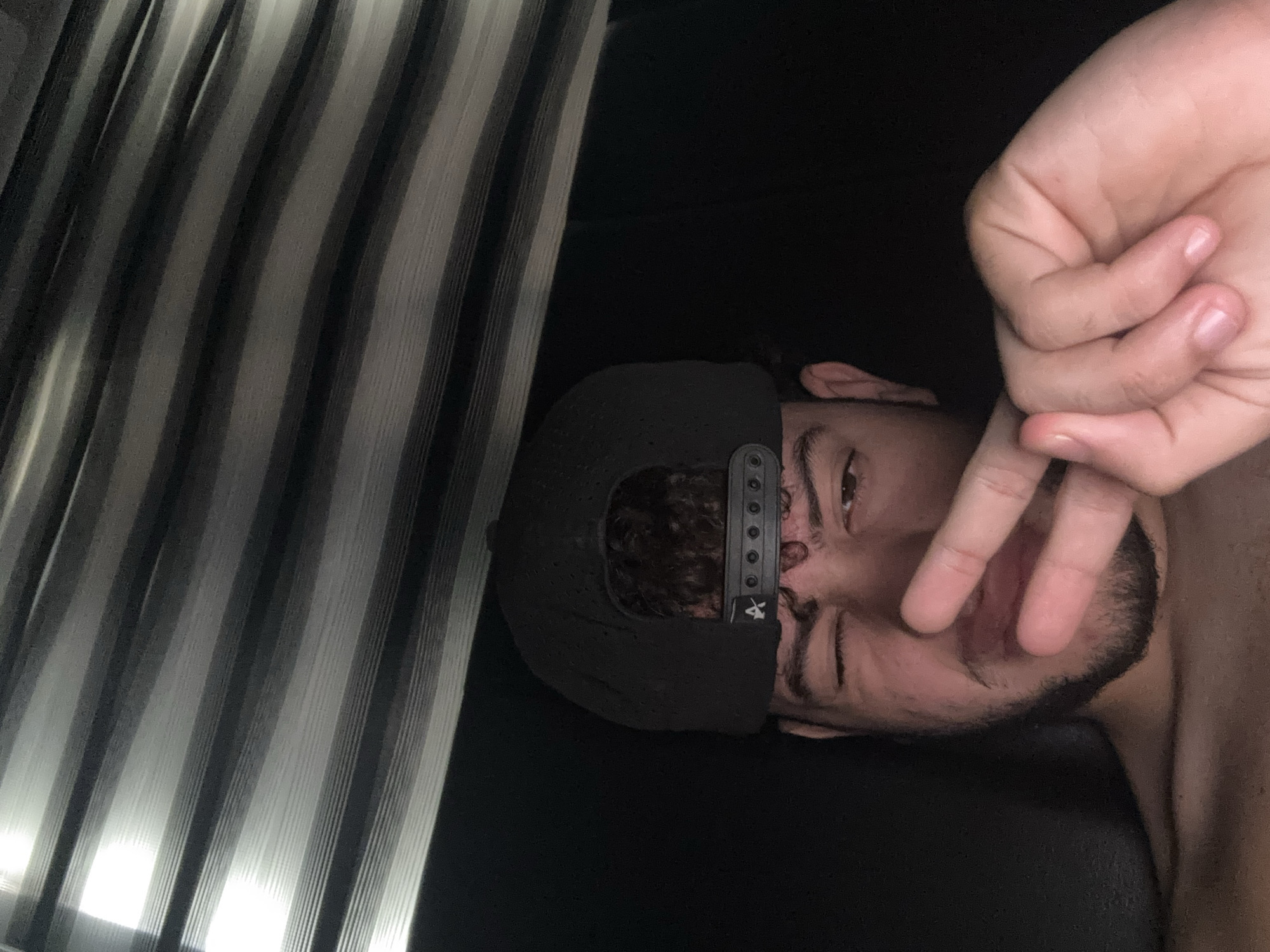 A young male wearing a black baseball cap with a logo is pictured from the chest up, in a low-angle, close-up shot. He is looking directly at the camera with one eye closed in a wink. His right hand is raised in front of his face, forming a peace sign with his index and middle fingers. His skin tone is light brown, and he has curly brown hair peeking out from under his cap, along with a beard. The background consists of horizontal blinds, with a bright light source coming from the top left, casting shadows and creating a striped pattern. The overall lighting is dim, with a warm tone. The setting is indoors, likely a bedroom, given the presence of blinds and the intimate nature of the selfie. The location in the city is Unknown, Unknown.