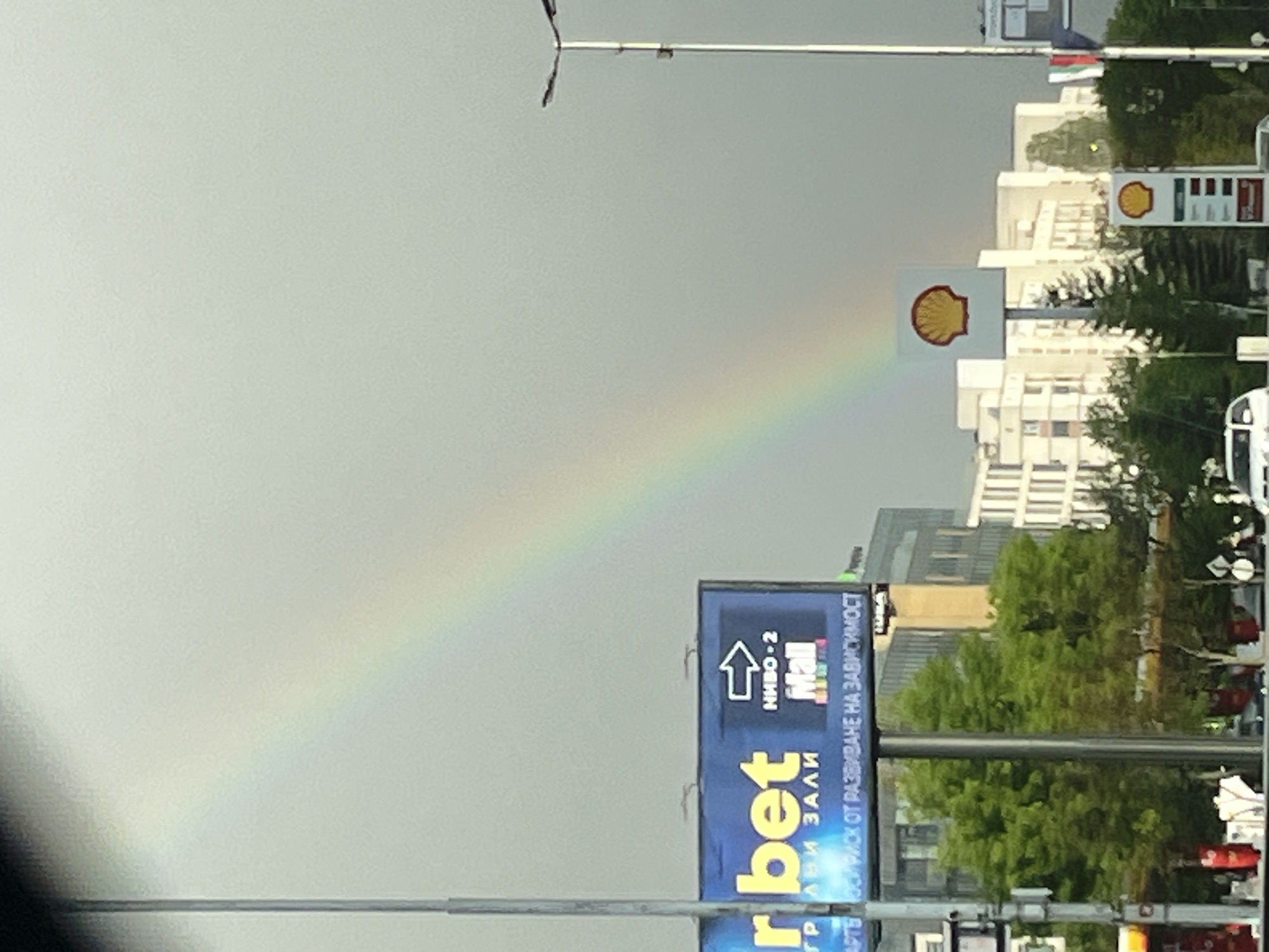 The image shows a partial rainbow in Sofia, Bulgaria. The rainbow arcs from the grey sky down toward a Shell gas station sign. In the foreground is an advertising billboard for "bet" and "Mall" in Cyrillic letters, as well as poles for traffic lights. Buildings and trees are visible in the background.