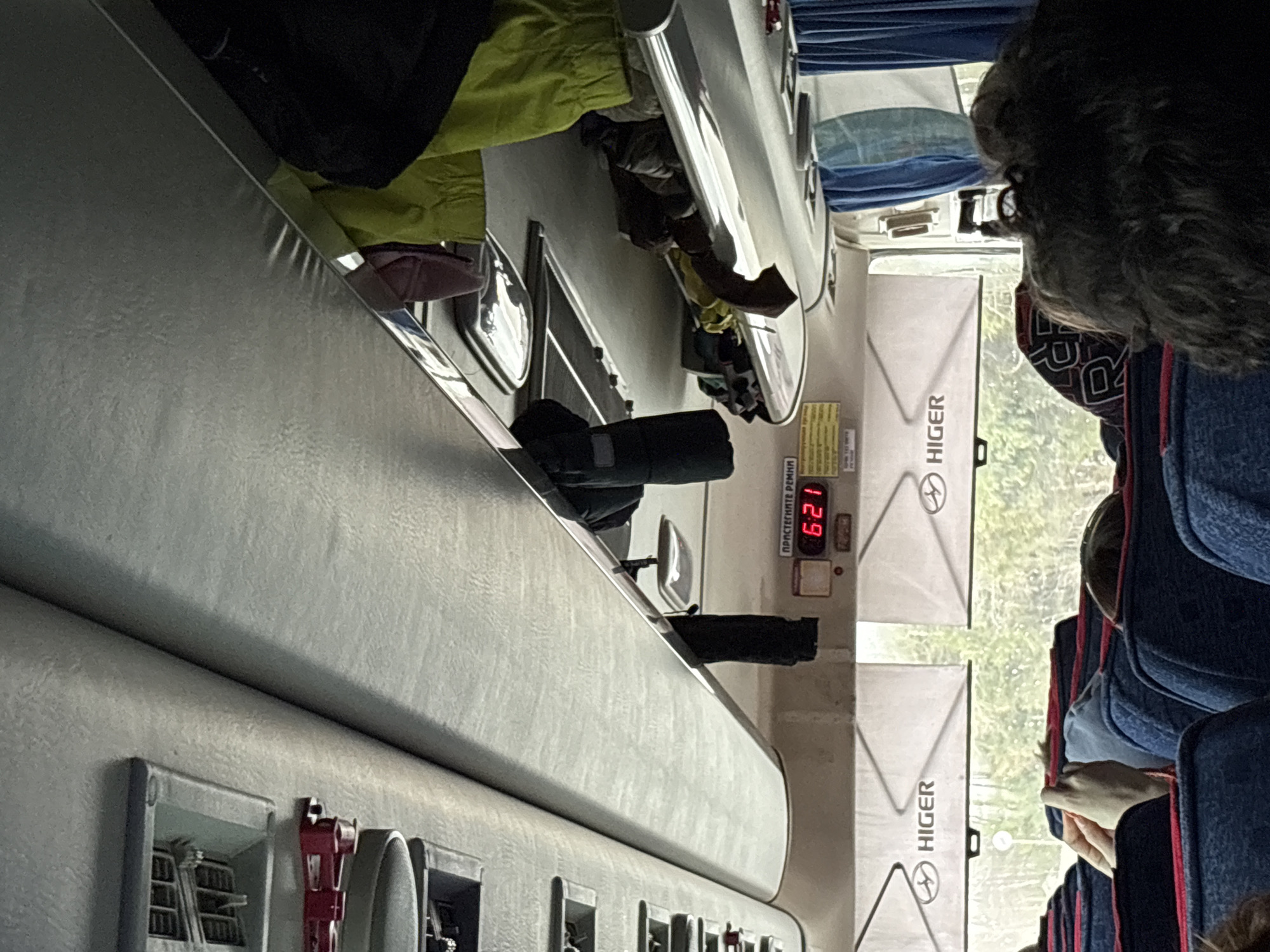 The image, taken inside a bus, shows an interior view with overhead storage, seats, and windows. The bus has a digital display showing "6:21" and branding for "HIGER" on the window panel. Passengers occupy the seats. There are indications of a city environment visible through the windows, which suggests the bus is moving. Based on the image metadata or any explicit indication, the location can not be definitively confirmed as Челва, Russia.