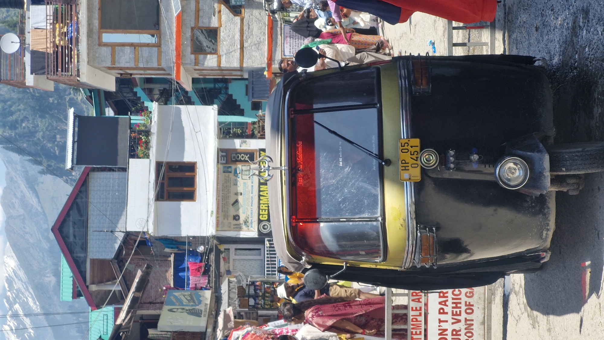 In Vashisht, India, an auto-rickshaw is parked on a street with buildings and mountains in the background. The auto-rickshaw is predominantly black and gold, with a yellow license plate reading "HP 05. 0451." A red inscription on the rear window says "Jai Maa Hadimba." Several people are visible in the background, including a woman in a pink sari on the left and a man in a white hat. Clothes are hanging on a balcony of a building on the left. The environment suggests a bustling street scene in a mountainous region.