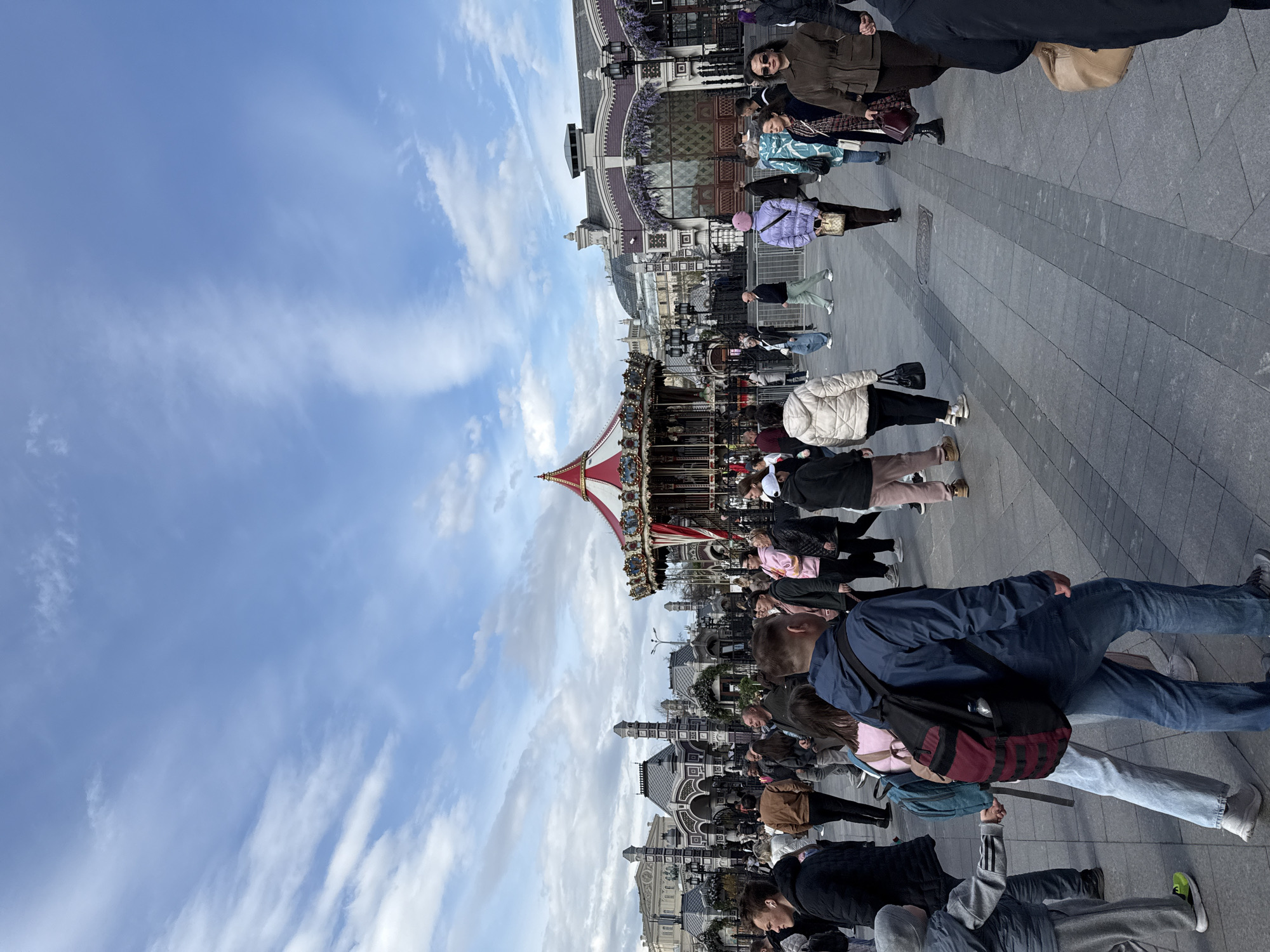 The image shows a crowded square in Moscow, Russia. A traditional carousel with a red and white striped tent roof is in the center of the frame. Numerous people are walking in different directions on a gray stone-paved square. A building with ornate details and dark-framed windows is in the background, behind the carousel. A pale blue sky with scattered white clouds is above the buildings and carousel.