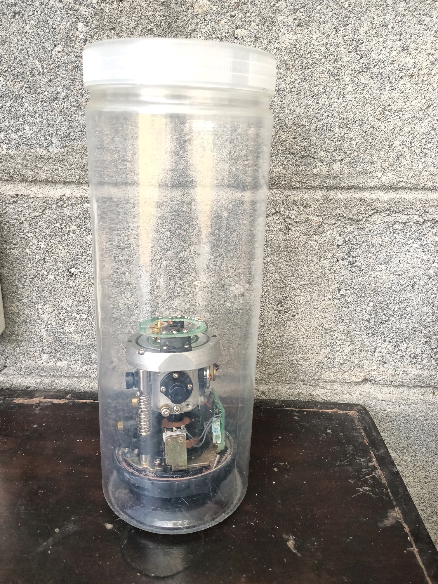 A clear, cylindrical plastic container with a white lid stands upright on a dark, wooden surface. Inside the container, a complex mechanical device with visible circuit boards, wires, springs, and metallic components is housed. The container is positioned in front of a rough, textured gray concrete wall. The scene appears to be outdoors, possibly in an industrial or workshop setting. The specific location within the city of Trenggalek, Indonesia, cannot be determined from the image alone.