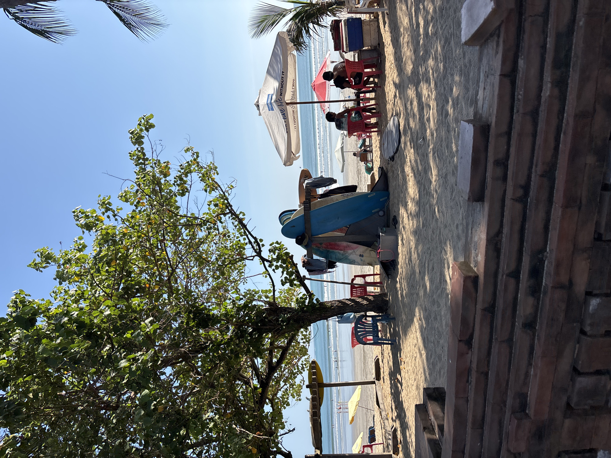 A bright, sunny day at Kuta Beach, Indonesia. The foreground is dominated by a brick structure, possibly a retaining wall or tiered seating, leading onto a sandy beach. A large tree with lush green foliage provides shade in the mid-ground, with its branches extending across the frame. Beneath the tree, a collection of surfboards is propped up, some leaning against a wooden rack. Several plastic chairs are scattered on the sand, with one blue chair positioned prominently in front of the surfboards. A red cooler and a grey plastic bin are also visible.
Further back, the beach stretches towards the ocean, with gentle waves rolling in. Several beach umbrellas, including yellow and red ones, are set up. In the distance, a few people are visible on the beach and in the water, some appear to be surfing or wading. Underneath a large white umbrella with text, two men are seated at a table, facing away from the camera. A smaller red umbrella is also present. The sky is a clear, vibrant blue. Palm fronds are visible in the upper right corner of the image. The overall scene depicts a relaxed beach atmosphere with a focus on surfing culture.