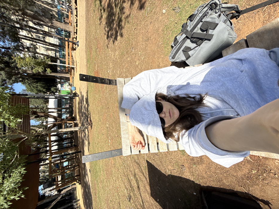 A person is depicted in a high-angle selfie photograph, lying supine on a slatted wooden bench with black metal supports. The individual wears a light gray hooded sweatshirt with the hood pulled over their head, dark sunglasses, and light-colored trousers. Their left arm is extended towards the camera, while their right arm rests on the back of the bench. A gray bag with black straps is positioned on the bench to the person's right.
The scene is an urban park in Ramat Gan, Israel, under bright daylight. The ground is dry, reddish-brown earth with sparse green grass and scattered leaf litter, exhibiting tree shadows. In the background, numerous green-leafed trees are visible. To the left, a wooden playground structure with dark metal framing stands. Further back, residential buildings are partially discernible through the trees. A light blue ball rests on the ground in the mid-background, and additional wooden park structures are present further to the right. The sky is clear blue.