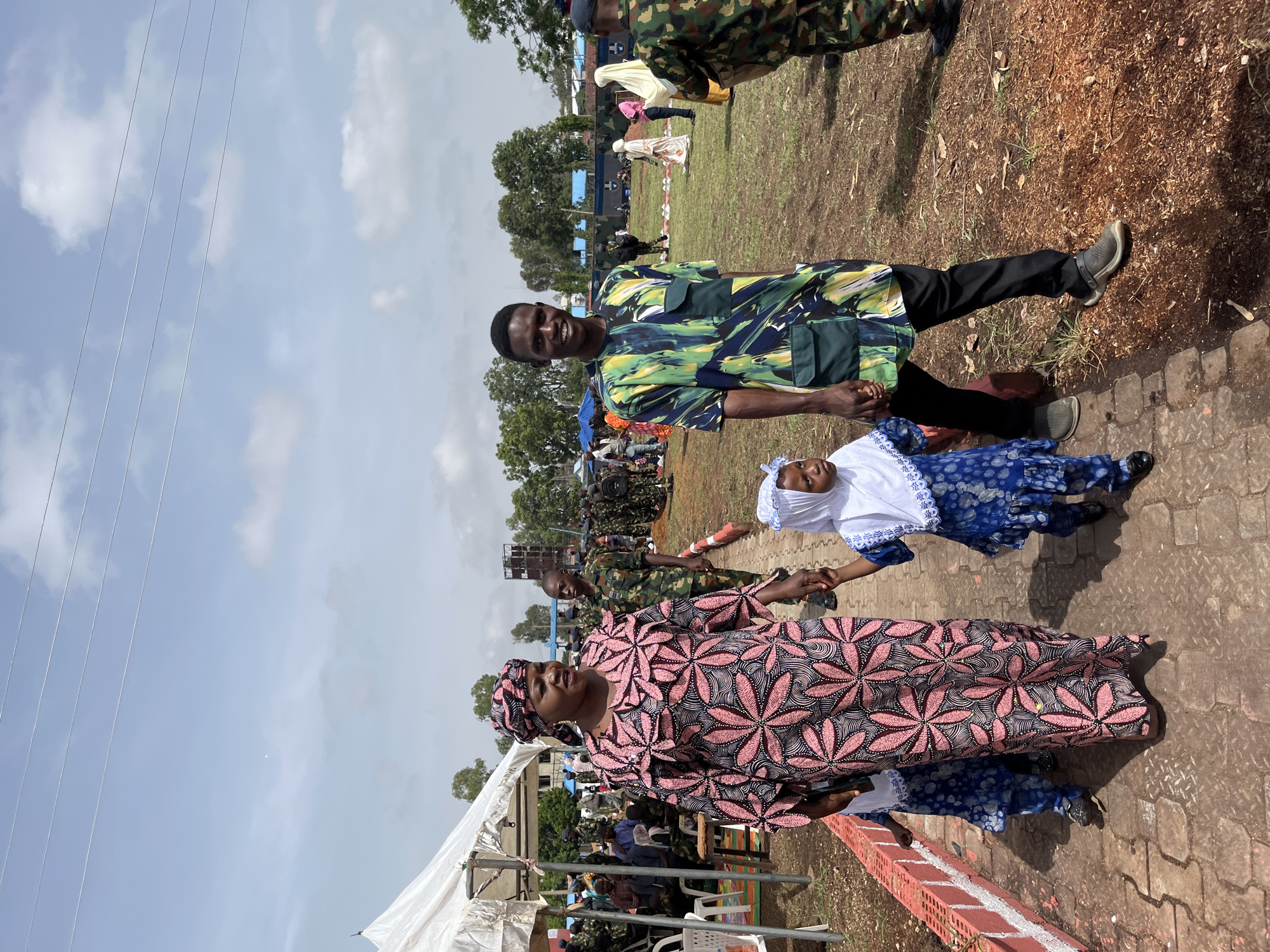 A family is walking on a paved path at an outdoor event in Barakin Lerawi, Nigeria. A woman in a pink and black patterned dress and headscarf walks in the foreground, holding the hand of a young girl in a blue and white dress and headscarf. Next to them, a young man in a colorful green, blue, and yellow patterned shirt and black pants smiles at the camera, also holding the girl's hand. Behind them, several people in military uniforms are visible, as well as tents, chairs, and other attendees scattered across a grassy field. The sky is bright blue with scattered clouds.