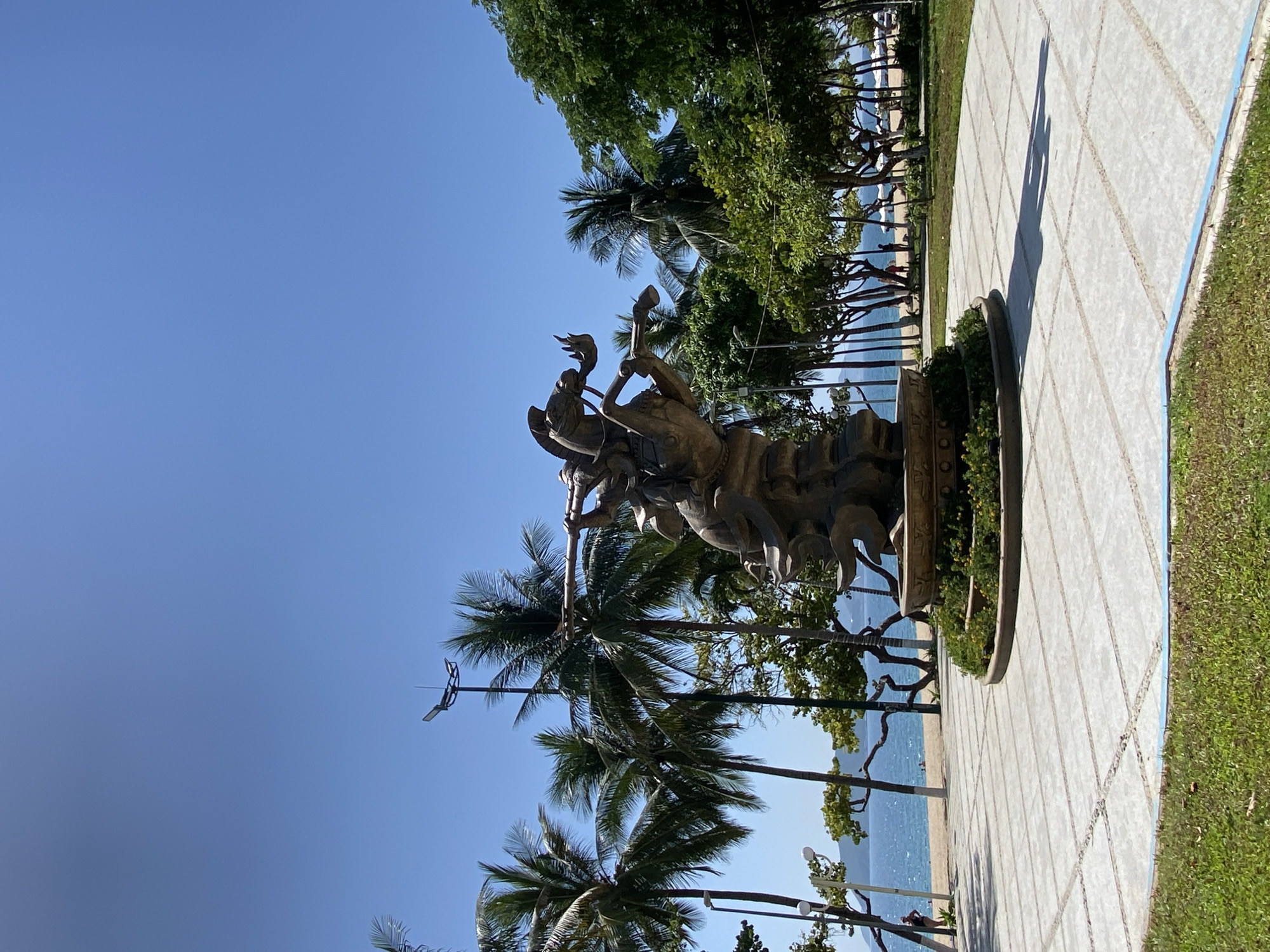 This is a photograph of a bronze statue of a warrior on horseback, located in a paved public area near the beach in Nha Trang, Vietnam. The statue is the central focus of the image, depicted in a dynamic pose, with the warrior holding a spear. The statue is situated on a circular base surrounded by green foliage.
Behind the statue, palm trees and other lush green trees line the area, with the blue ocean and a sandy beach visible in the background. The sky is clear and blue. Several people are seen on the beach, some walking and some relaxing. A paved walkway extends from the statue area towards the beach. Streetlights are also visible in the background among the palm trees. The overall scene depicts a sunny day in a scenic coastal city.