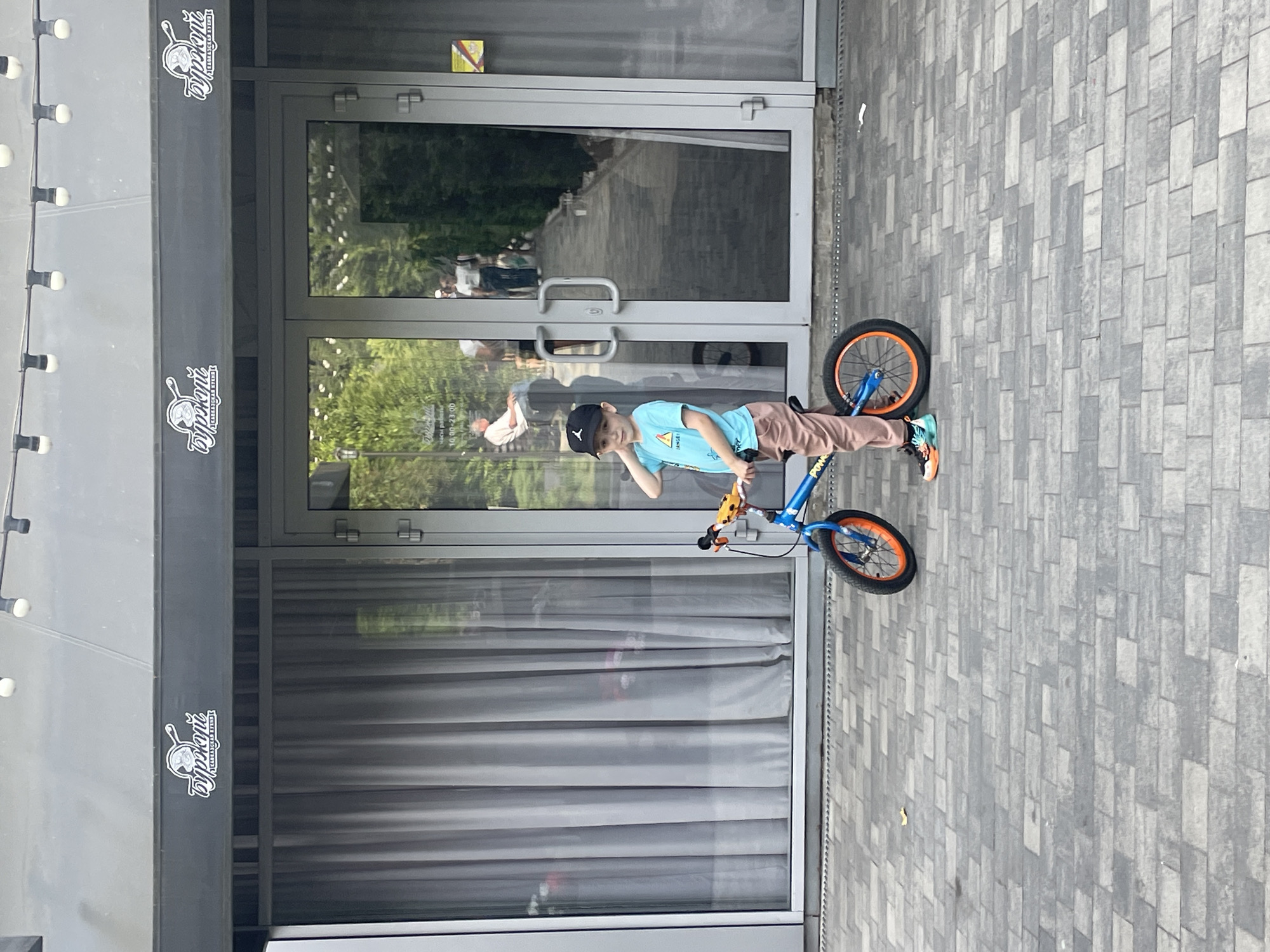 A young boy, wearing a black baseball cap, a light blue t-shirt, and light brown pants, stands on a paved walkway in front of a modern building with large glass doors and windows. He is posing with a blue and orange balance bike. The building appears to be a cafe or restaurant, as indicated by the signage featuring the word "БурЖуи" and "КАВКАЗСКАЯ КУХНЯ" (Caucasian Cuisine), and hours of operation "10.00 - 23.00". The walkway is made of grey rectangular paving stones. In the background, reflections in the glass reveal trees and people walking. The lighting suggests it is daytime. The location is identified as Sochi, Russia.