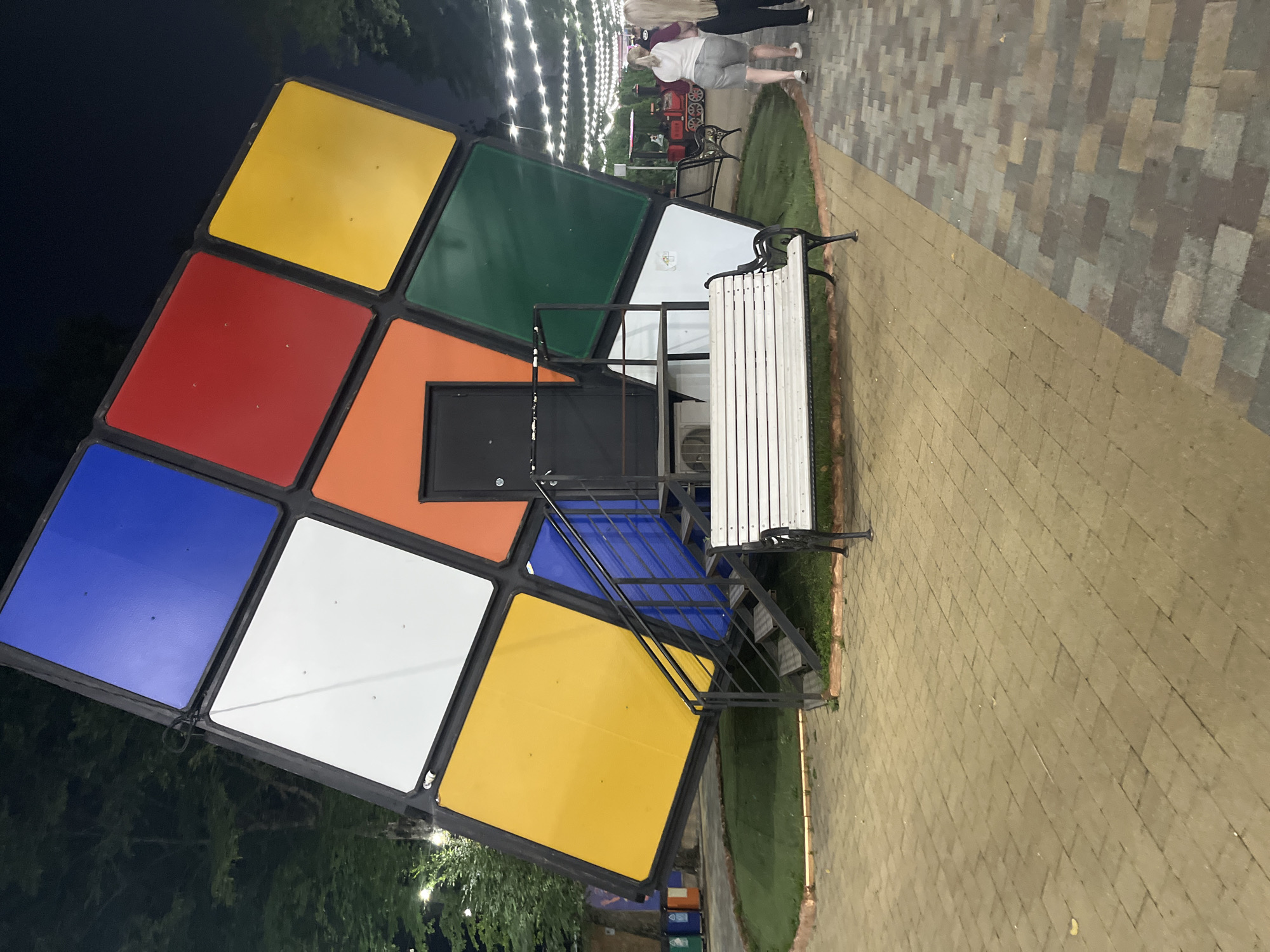 An outdoor, nighttime scene captures a large, tilted Rubik's Cube structure with a dark doorway and metal stairs leading to it. In front of the cube, a white wooden bench with black metal legs sits on a paved walkway. To the right of the bench, two women are walking away from the camera on a different section of the paved walkway, which is illuminated by overhead string lights. Further to the right, a miniature train with people aboard is visible. The background features trees and more string lights. The Rubik's Cube itself is composed of colored panels, including blue, white, red, yellow, orange, and green. The location is identified as the park of the Victory, Stavropol, Russia.