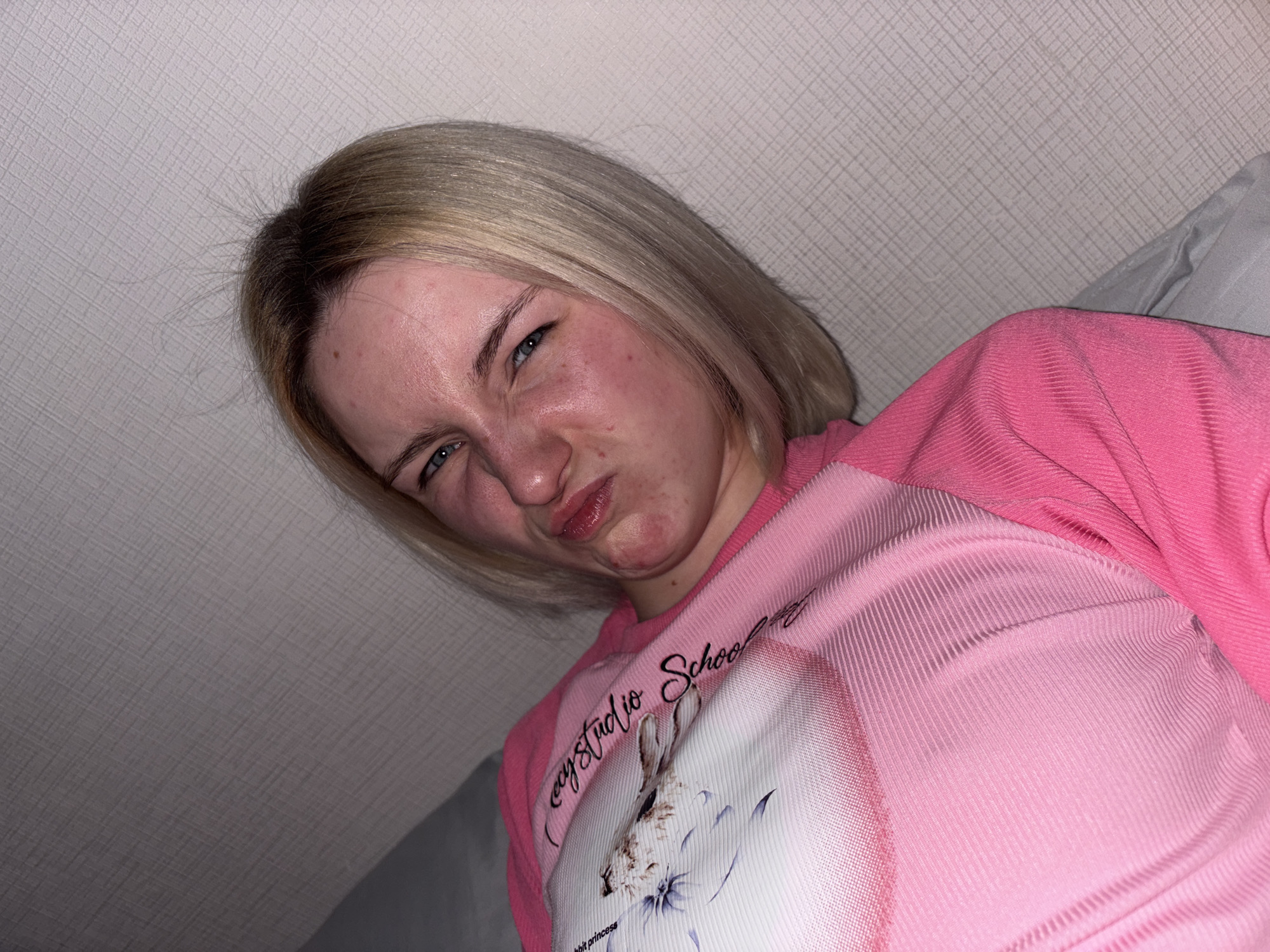 An indoor, close-up photograph features a young woman with shoulder-length blonde hair and darker roots, making a playful or exaggerated "disgusted" expression. Her eyes are squinted, nose wrinkled, and lips pursed. Her cheeks show a slight flush.
She is wearing a long-sleeved pink top. The sleeves and upper part of the shirt are made of a ribbed, textured fabric. The central panel of the shirt is a smoother, lighter pink, featuring a graphic print of a light-colored rabbit surrounded by delicate blue and purple floral designs. Above the rabbit, cursive text reads "cozystudio School," and below, partially visible text says "abbit princess."
The background consists of a light-colored wall with a subtle, textured geometric pattern, possibly wallpaper. To the right of the subject, a soft gray fabric, likely a pillow or bedding, is visible. The lighting suggests an indoor setting, likely artificial. The woman is angled, with the shot taken from a slightly low perspective. The scene is set in Ligolambi, Russia.