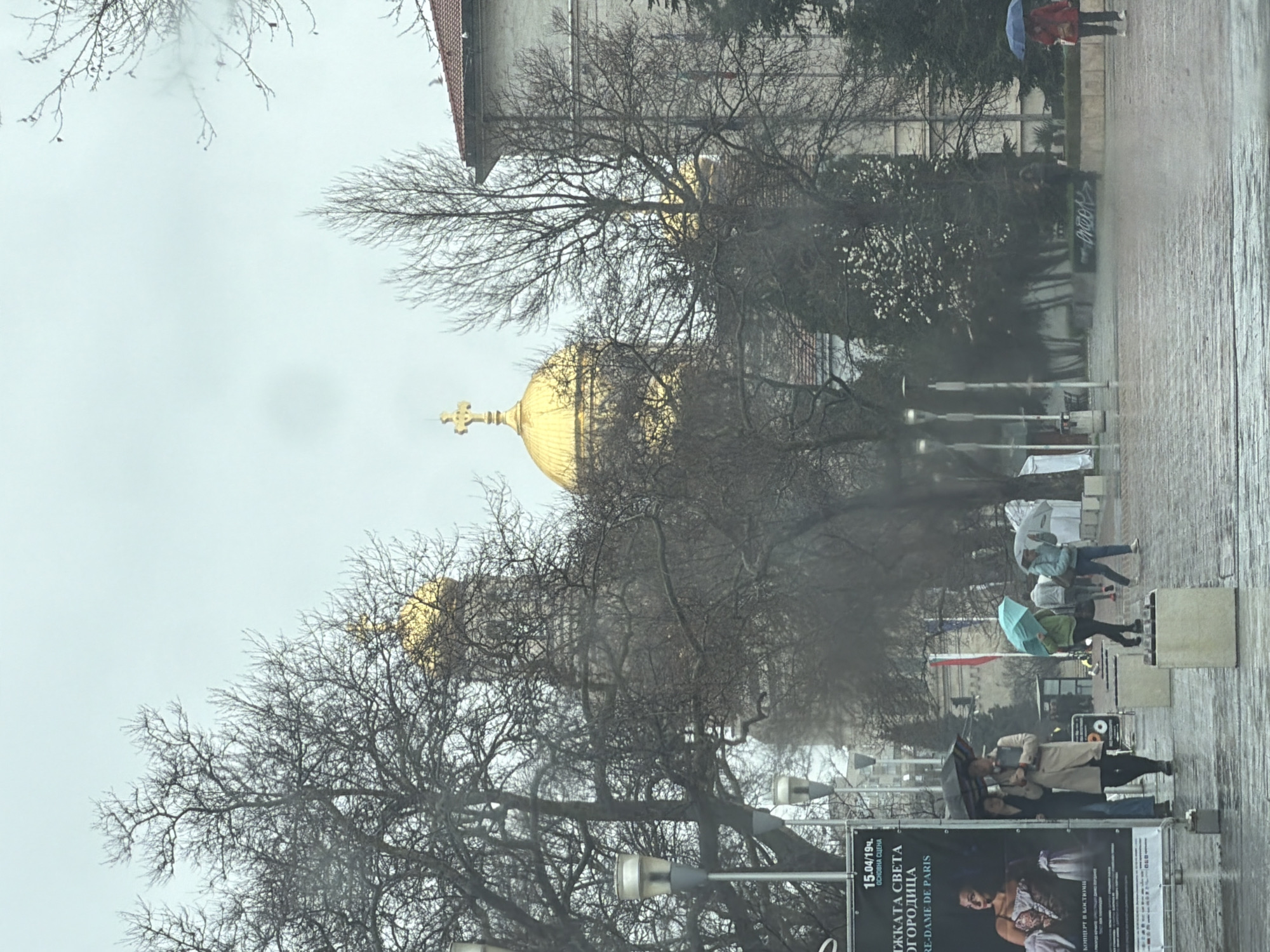 The image shows a rainy day in Varna, Bulgaria, with a clear view of the golden domes of the Dormition of the Mother of God Cathedral. Bare trees frame the domes, adding to the somber mood of the scene. In the foreground, a street scene unfolds with people walking along the sidewalk, many holding umbrellas against the rain. A large poster is visible at the bottom, advertising a concert. The light is flat and diffused due to the overcast sky, enhancing the damp and cool atmosphere of the day.