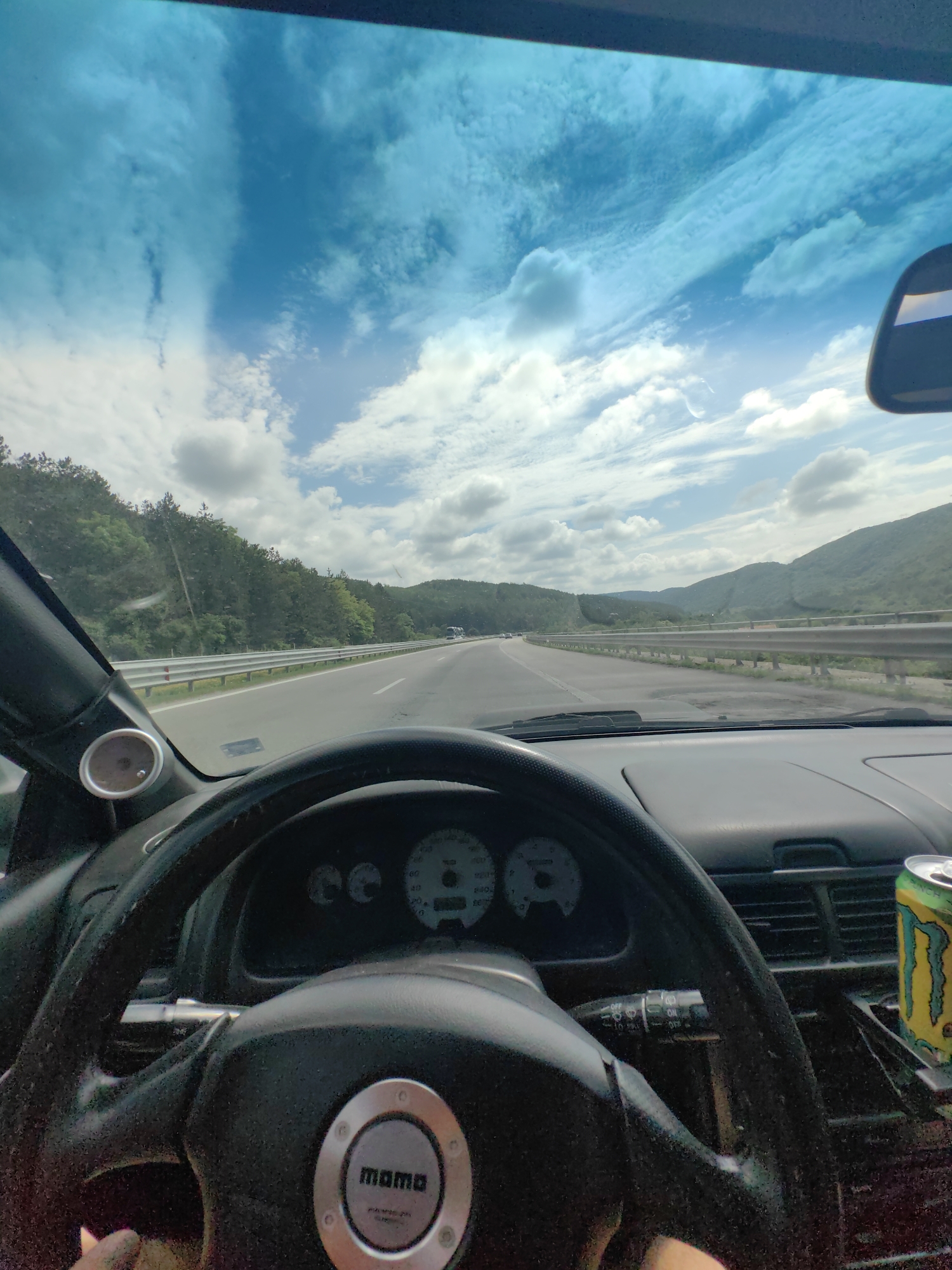 The image is a first-person perspective from inside a car, looking forward through the windshield. The car is on a highway, with guardrails lining the right side and a dense forest on a hillside to the left. The sky is blue with scattered white clouds. The dashboard of the car is visible, including the steering wheel, instrument cluster, and a can of Monster Energy drink. The steering wheel has a "momo" logo in the center. Based on the presence of the highway and the surrounding natural landscape, the scene is likely on a highway in or near Potop, Bulgaria.