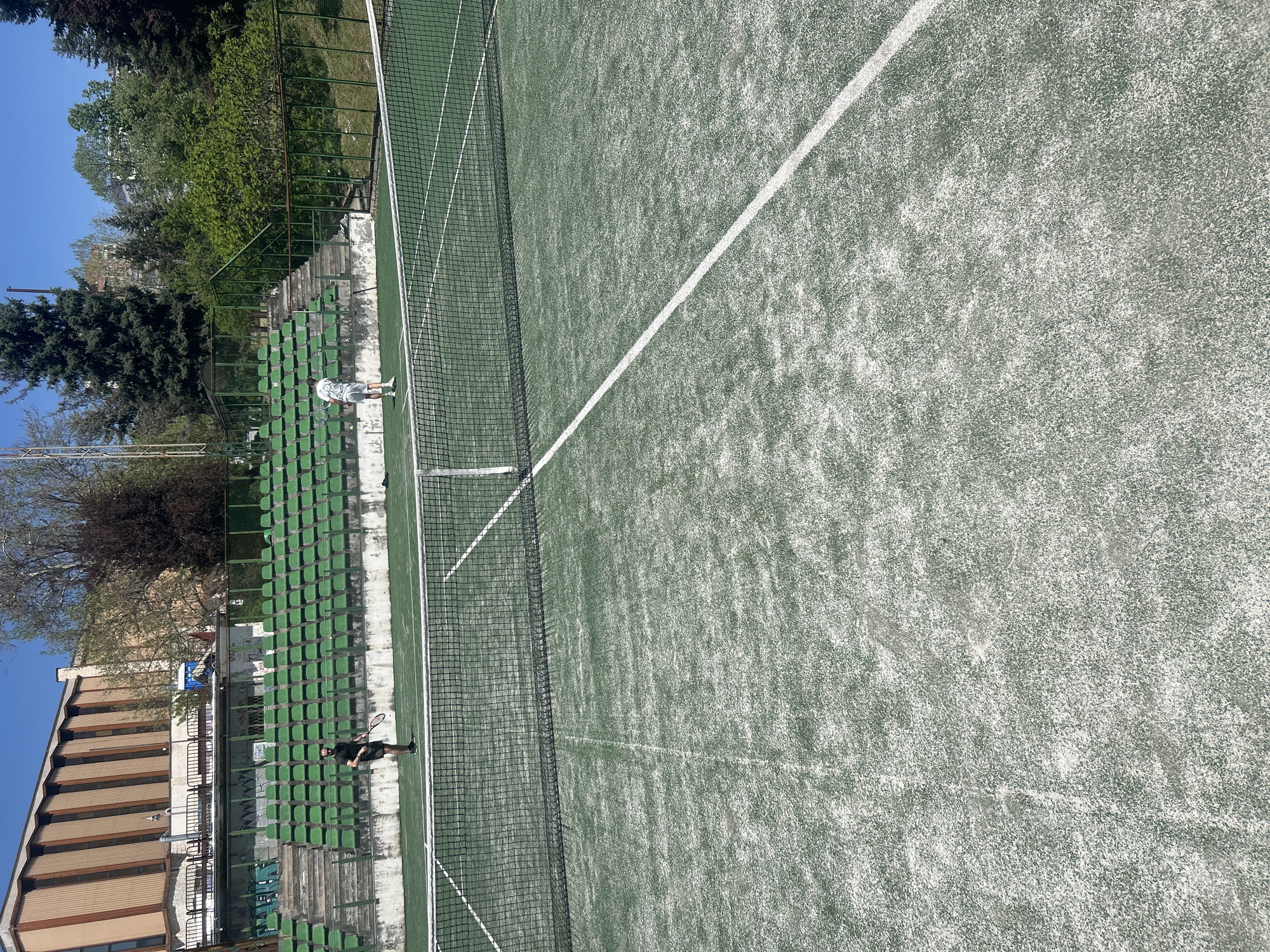 This image captures an outdoor tennis court in Veliko Tarnovo, Bulgaria, bathed in bright daylight under a clear blue sky. The court features a green playing surface with prominent white boundary lines and a black tennis net stretched across its middle. The court surface shows visible texture and some wear.
Two individuals, presumably tennis players, are present on the court, both holding rackets. One player, wearing a white top and shorts, stands in the mid-ground on one side of the net, near the spectator area. The second player, dressed in darker clothing, is positioned closer to the foreground and the net on the opposite side.
Along one side of the court, a long stretch of empty green stadium-style seating is visible, suggesting a sports facility. Beyond the seating, a diverse array of lush green trees, including both evergreens and deciduous types, forms a natural backdrop. To the left, a multi-story building with a brown roof is partially visible, likely part of a larger sports complex or adjacent infrastructure. The clear weather and empty stands indicate a casual setting, possibly a practice session or an informal game. No discernible text is visible.