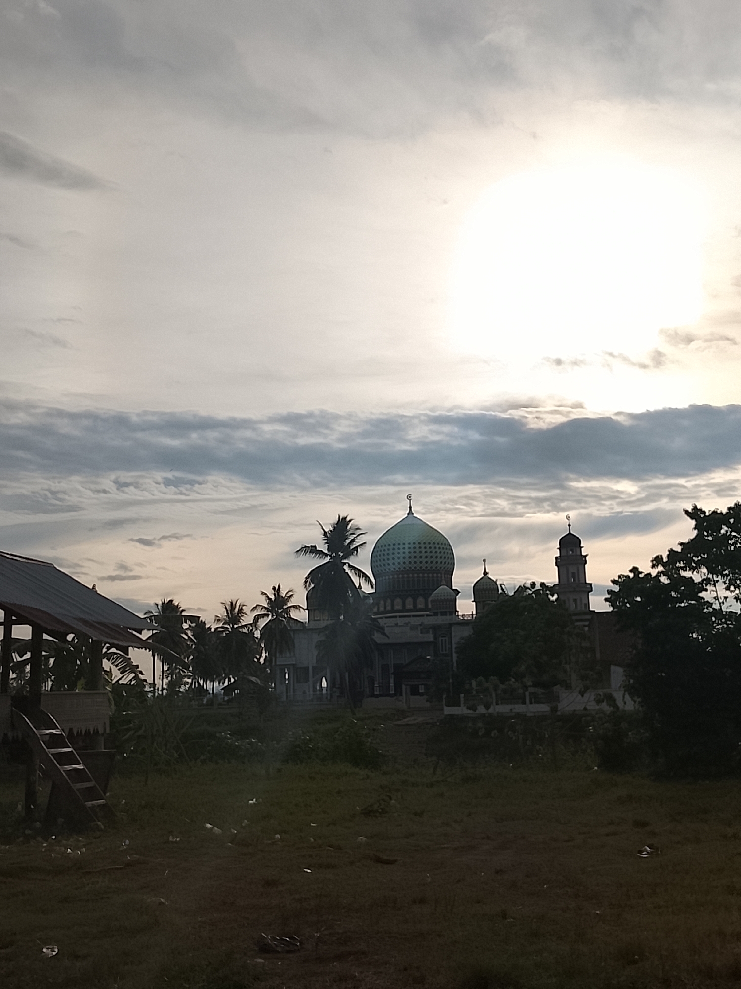 The image captures a mosque with a distinctive green dome, a minaret, and several smaller domes. The mosque is surrounded by trees and appears to be situated in a tropical or subtropical region, given the presence of palm trees. The sky is partly cloudy, with a bright, hazy sun visible in the upper right quadrant. In the foreground, there is a structure with a tin roof and a wooden ladder, possibly a small building or shelter. The ground in the foreground is covered in grass and some scattered debris. The overall impression is of a scene in Geunteng, Indonesia, possibly during the daytime, with an overcast sky diffused by sunlight. There are no people visible in the image.