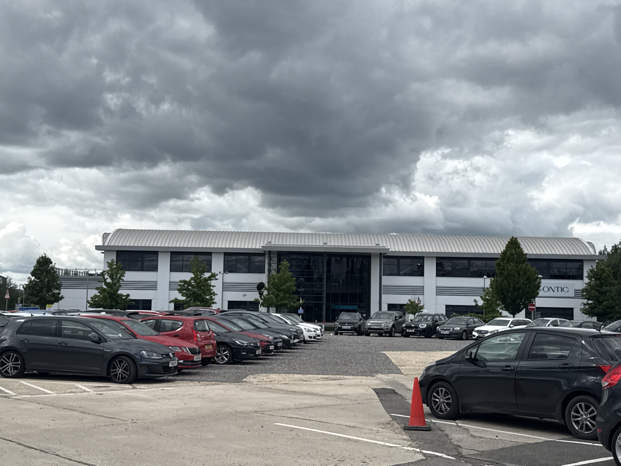 The image shows a parking lot with several cars parked in front of a modern office building. The building has a white facade with large windows and a curved roof. A sign on the building reads "SONTIC". The parking lot is paved and has white lines marking the parking spaces. There is a red traffic cone in the foreground. The sky is cloudy and dark, suggesting impending rain. The scene appears to be a typical office park environment on an overcast day. The location is Tewkesbury, United Kingdom.