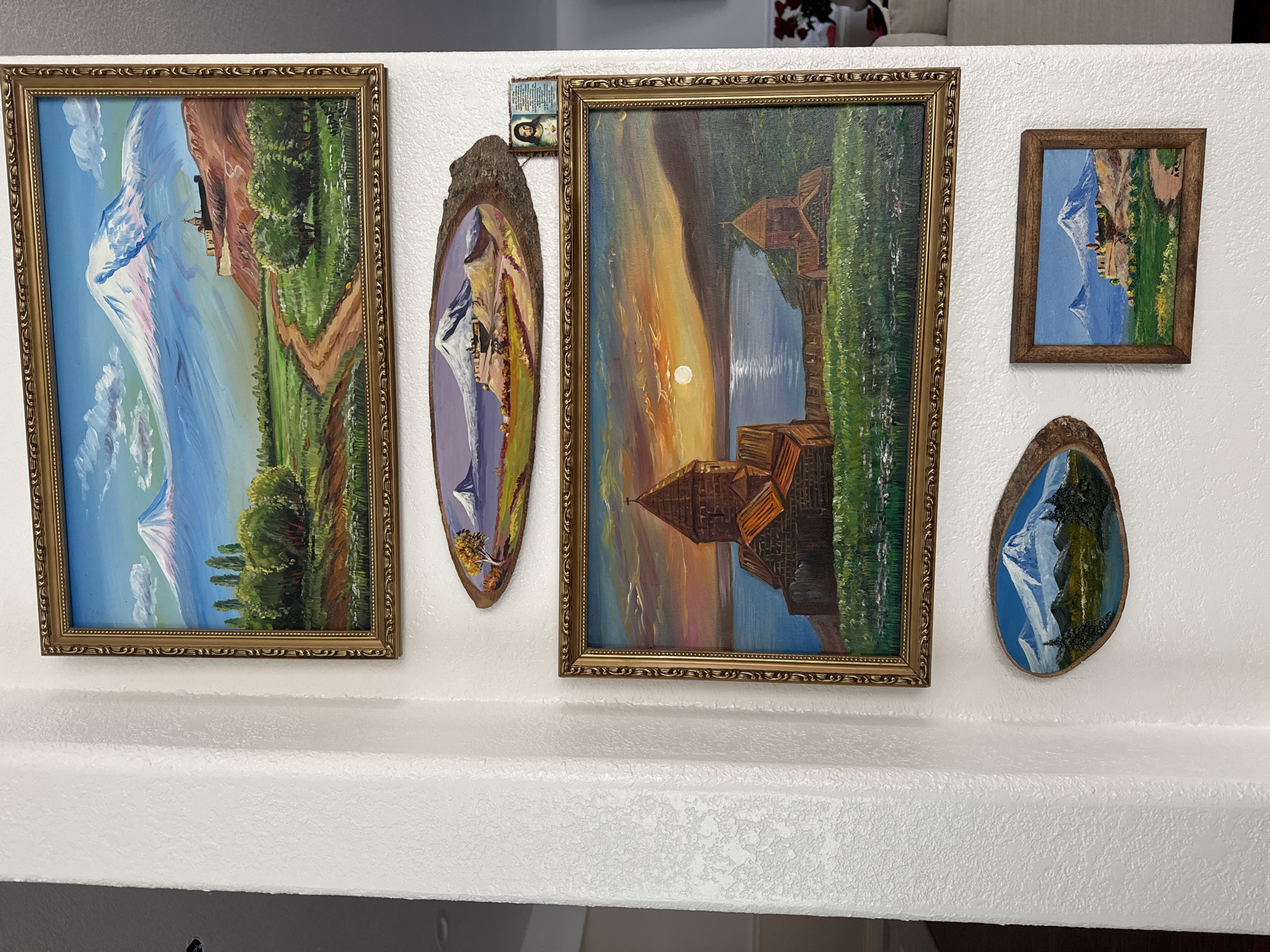 The image shows a wall in a room, likely a home, decorated with several paintings and a small religious icon. The wall is painted white and has a textured finish. The paintings are of landscapes, predominantly featuring mountains, possibly Mount Ararat.
There are five framed artworks:
1. A large rectangular painting at the top features a majestic snow-capped mountain under a blue sky with scattered clouds. In the foreground, there is a green landscape with trees and a dirt path leading towards the mountain. The painting is in an ornate gold frame.
2. An oval-shaped painting, mounted on a piece of wood, depicts a similar mountain landscape with a body of water in the foreground and a structure on a hill.
3. A medium-sized rectangular painting, also in a gold frame, shows an old stone church with multiple domes by a body of water during a sunset. The sky is filled with vibrant orange, yellow, and purple hues. Green grassy fields surround the church.
4. A small, oval-shaped painting, similar in style to the second painting, displays a snow-capped mountain and evergreen trees reflected in a lake.
5. A small rectangular painting in a dark wood frame shows a mountain range with a castle-like structure on a hill, set against a blue sky with green fields in the foreground.
To the right of the oval wooden painting (number 2), there is a small religious icon or framed image of Jesus Christ.
Based on the provided metadata, the location of this image is in Clovis, United States of America. The scene appears to be an indoor setting, likely a living room or hallway, given the display of art on the wall. No people are visible in the image, and there are no specific actions or interactions taking place. The environment is clean and well-lit, with the focus being on the art collection.