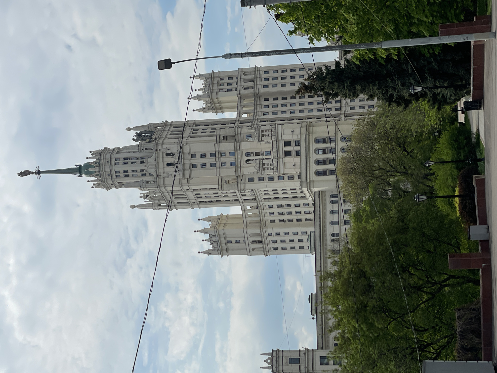 The image shows the Ministry of Foreign Affairs building in Moscow, Russia, a prominent Stalinist skyscraper. The building is white with intricate architectural details and a spire topped with a Soviet star. It is viewed from a low angle, emphasizing its height. The foreground includes trees, lampposts, and power lines, while the background consists of a cloudy sky. The environment is urban, with hints of surrounding streets and sidewalks.