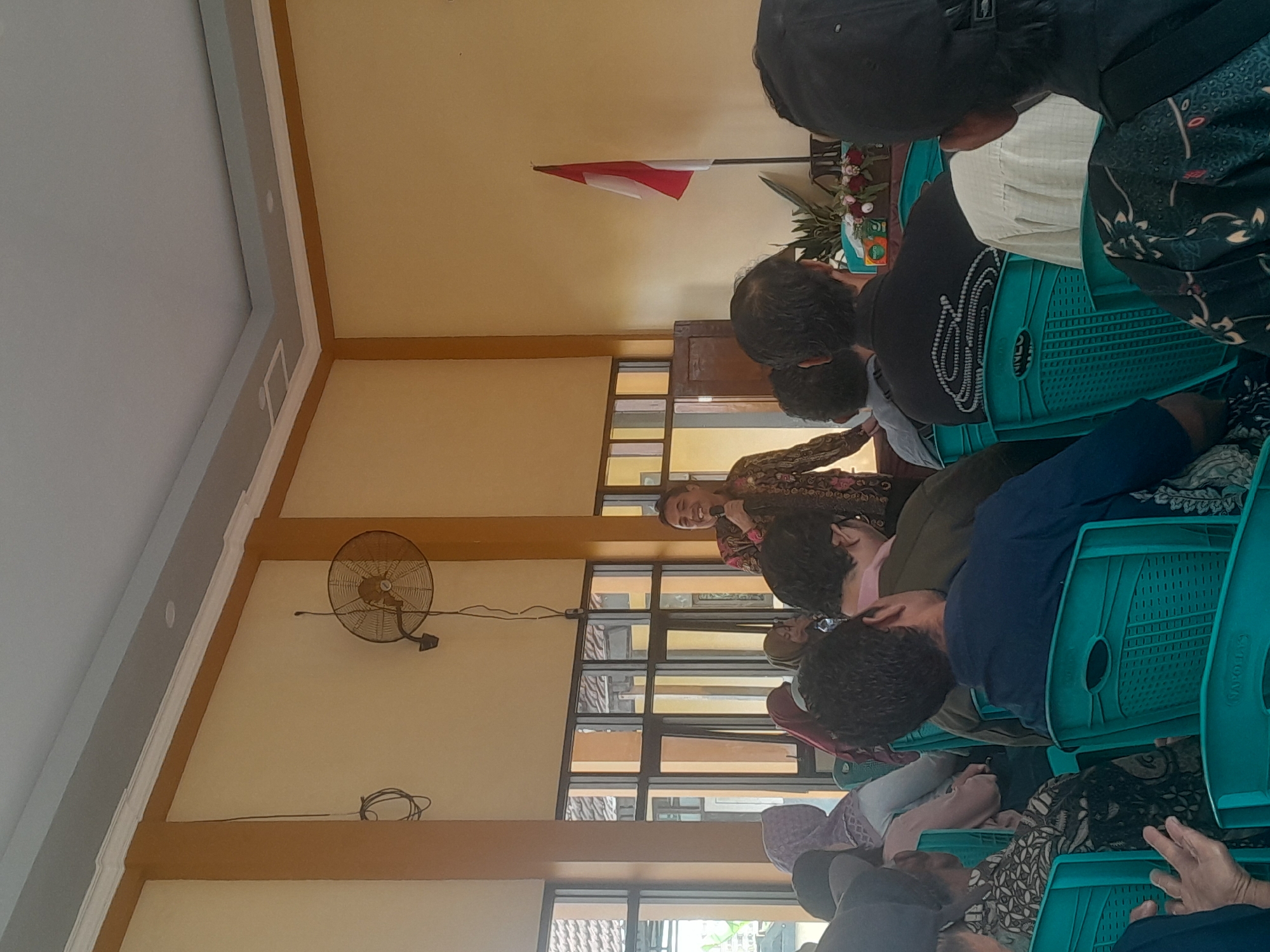 The image captures an indoor event, likely a seminar or meeting, held in Jajag, Indonesia. A man in traditional Indonesian batik clothing stands on the right side of the room, holding a microphone and addressing an audience seated on teal-colored chairs. The room has yellow walls with wooden accents and large windows that reveal glimpses of the outdoors. A wall-mounted fan is visible on the left side of the room, and an Indonesian flag stands on a pole near the back. The audience members are diverse, with some wearing headscarves. The overall atmosphere appears to be one of engagement and formal gathering.