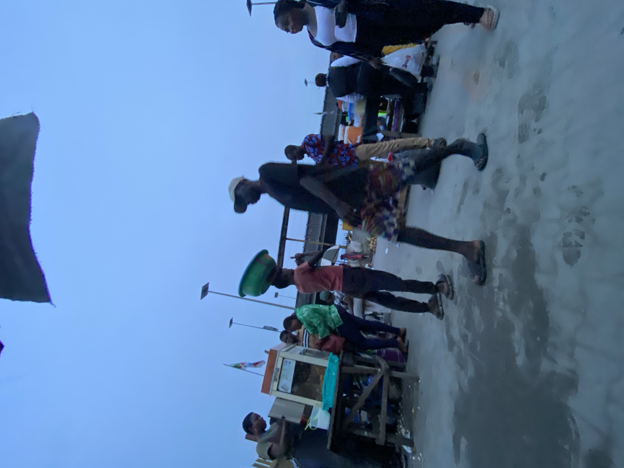 The image depicts an outdoor scene in Lekki, Nigeria, under an overcast sky. Several people are present, many appearing to be engaged in commerce or street vending. A man is carrying a green basin on his head, while others stand near a food stall. The ground is paved and appears slightly wet. In the background, there are streetlights and what may be a national flag.