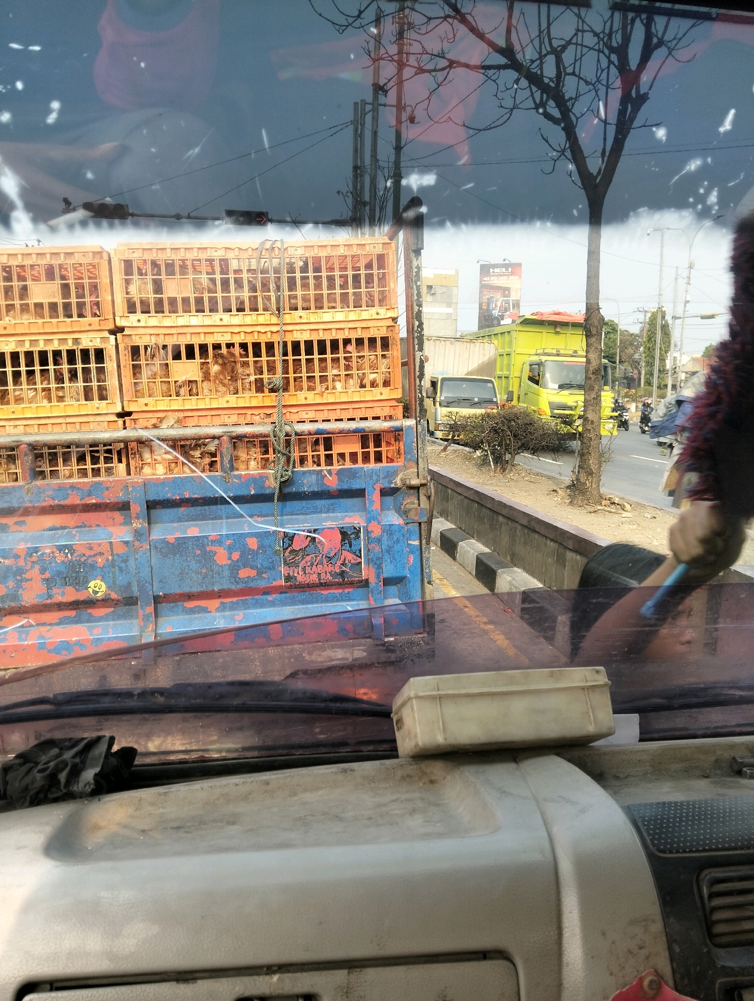 The image captures a street scene in Semarang, Indonesia, viewed from the interior of a vehicle. In the foreground, the dashboard of the vehicle is visible, with a white plastic container resting on it. The windshield displays reflections, including a blurred image of a person and overhead wires.
Beyond the windshield, a large blue truck with red accents and orange crates stacked on its bed is prominent. These crates appear to contain livestock, possibly chickens, as indicated by the visible feathers and shapes within. A rope is attached to the crates and hangs down. The truck's side displays a sticker with text and a graphic of a person.
In the background, several other vehicles are present on a paved road. A yellow dump truck is parked alongside a smaller yellow truck. Further down the road, more vehicles, including motorcycles, are in motion. Trees and utility poles line the street, with buildings and a billboard visible in the distance. The overall environment suggests an urban or suburban setting.