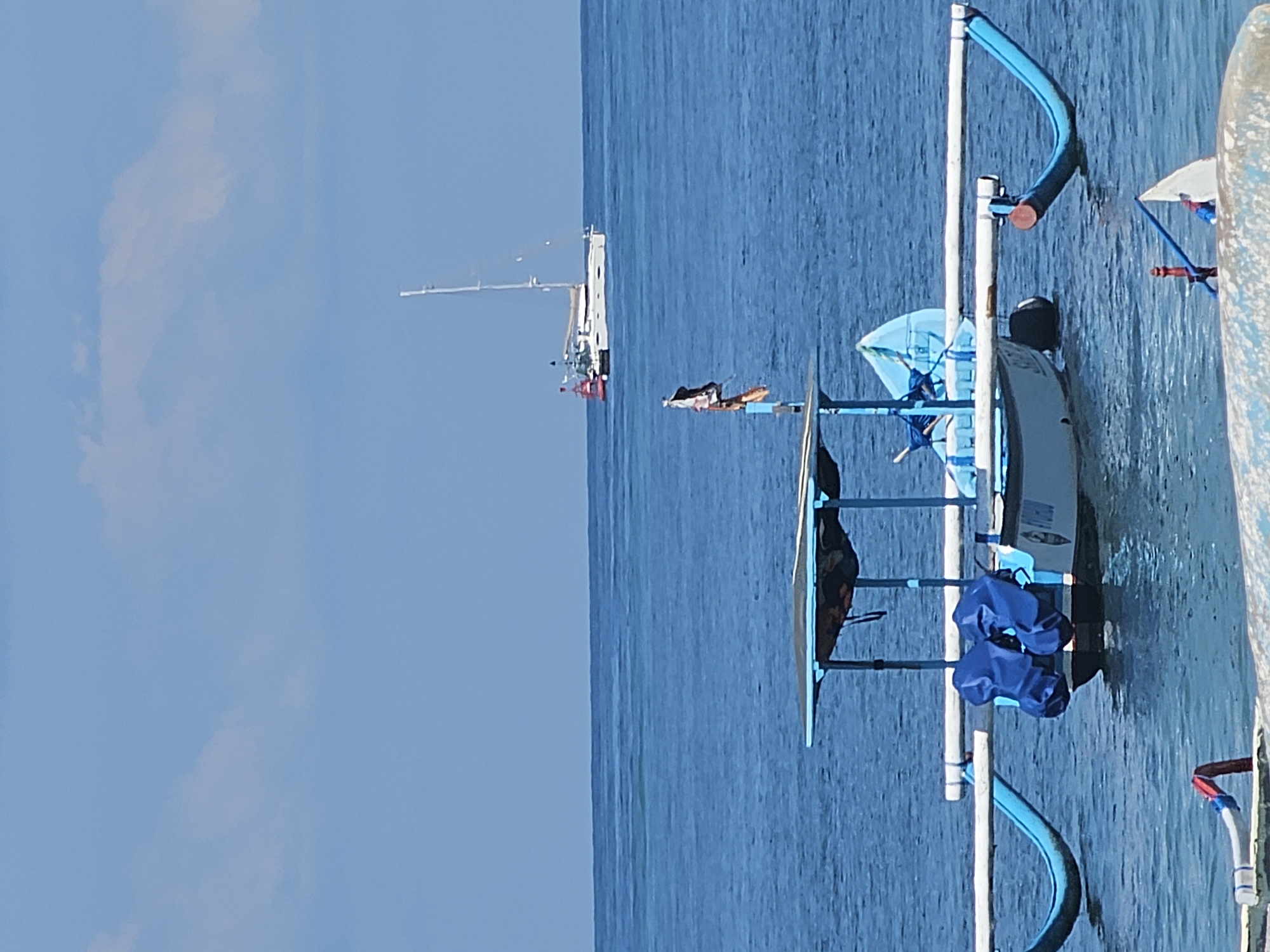 The image shows a boat on calm blue water under a clear blue sky. Another, larger boat is visible in the distance. The location is Denpasar, Indonesia.