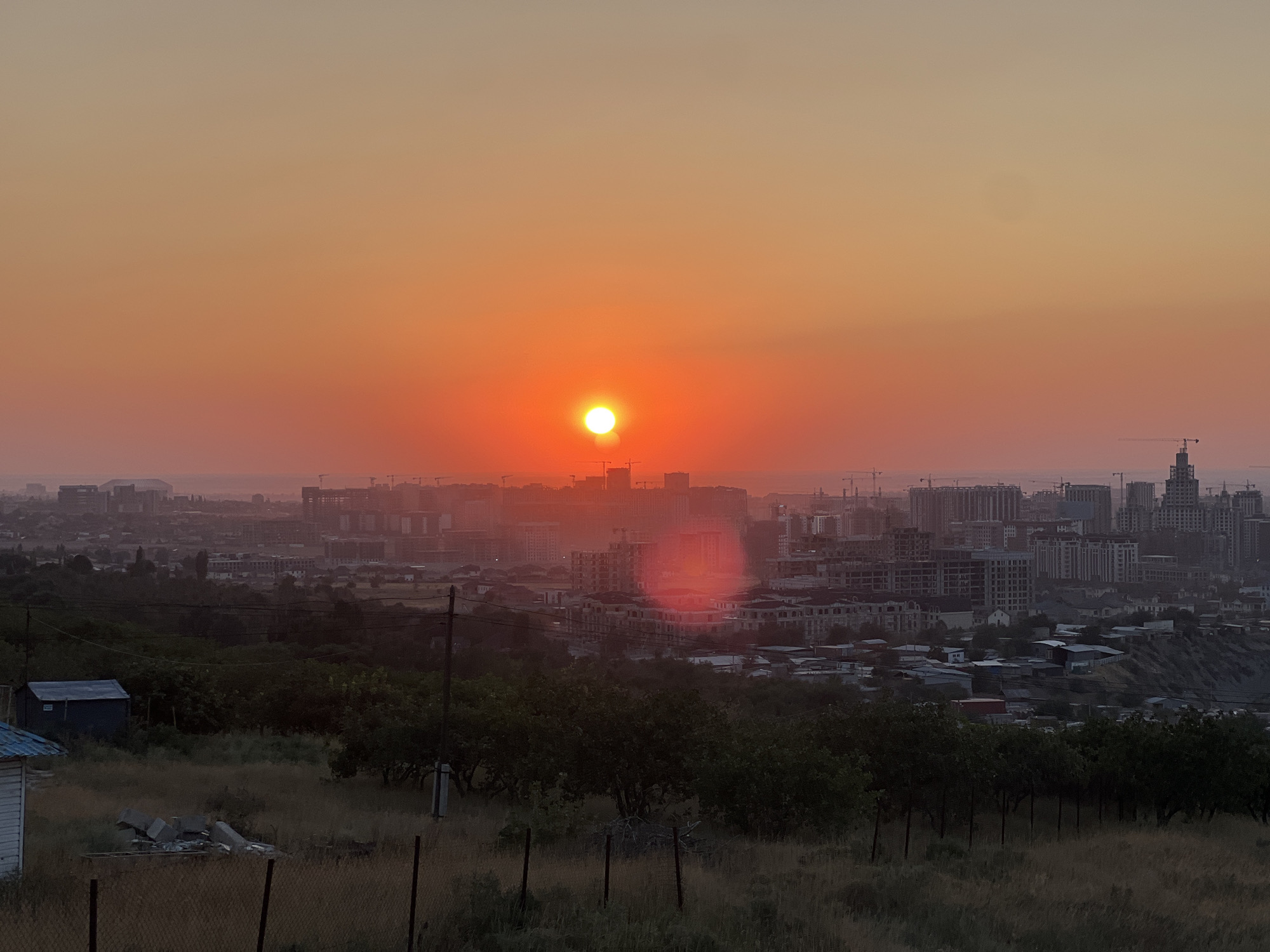 A wide-angle, outdoor, eye-level shot captures a sunset over Bishkek City, Kyrgyzstan. The sky is a gradient of orange and yellow, with the sun, a bright orange circle, positioned in the upper center. The cityscape stretches across the middle ground, with numerous buildings of varying heights, some appearing to be under construction with visible cranes. In the foreground, a grassy slope descends, dotted with dry grass and small trees. A section of a fence and a small, blue-roofed shed are visible on the left side of the frame. The overall impression is of a developing urban environment bathed in the warm hues of a setting sun.