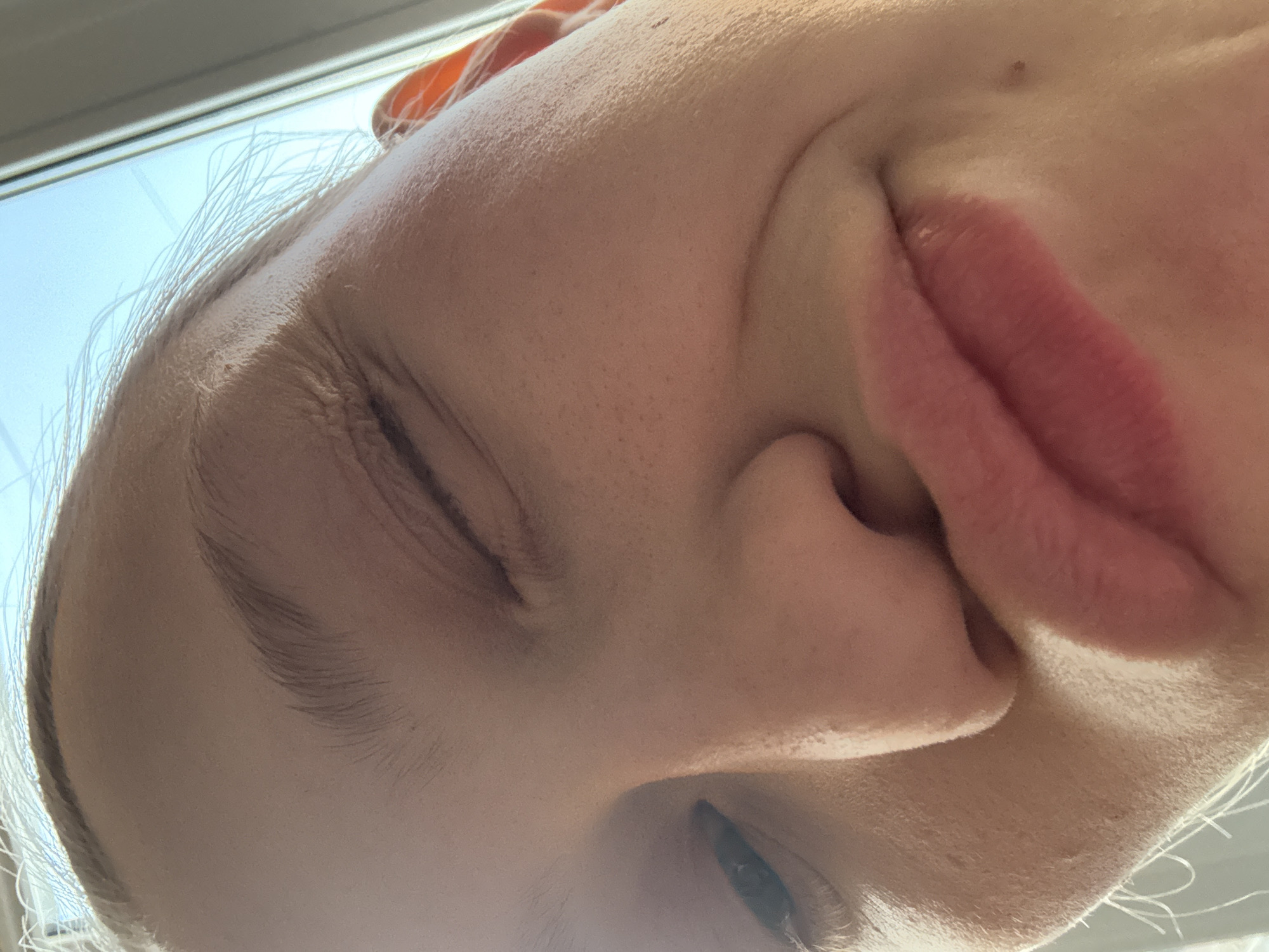 A close-up, tilted portrait of a fair-skinned person, possibly female, with one eye partially visible. The person has closed eyes, light eyebrows, and a small mole on their cheek. Their nose is slightly upturned, and their lips are full with a pink hue. Light-colored hair is visible around the face and near the window. The image appears to be taken indoors near a window, with bright light coming from outside. There is no indication that this image was taken in the city of Perm, Russia.