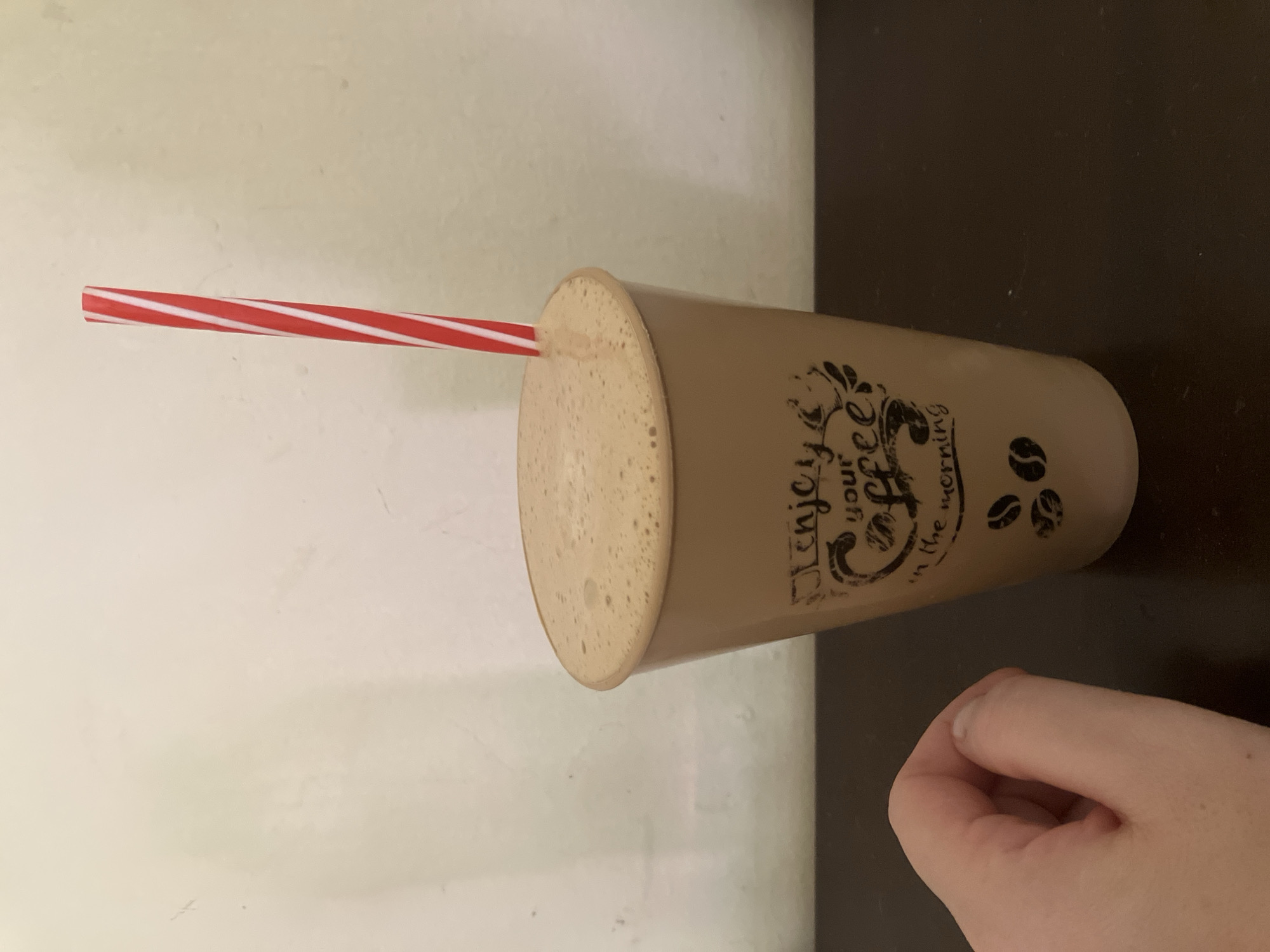 A tall, beige disposable cup with a foamy brown beverage is placed on a dark brown surface. A red and white striped straw emerges from the beverage. The cup has black text and graphics that read "Enjoy your Coffee in the morning" with two coffee beans at the bottom. A person's hand is visible on the left side of the image, resting on the surface. The background is a light-colored, textured wall. The image does not contain any information about the city of Paleo Faliro, Greece.